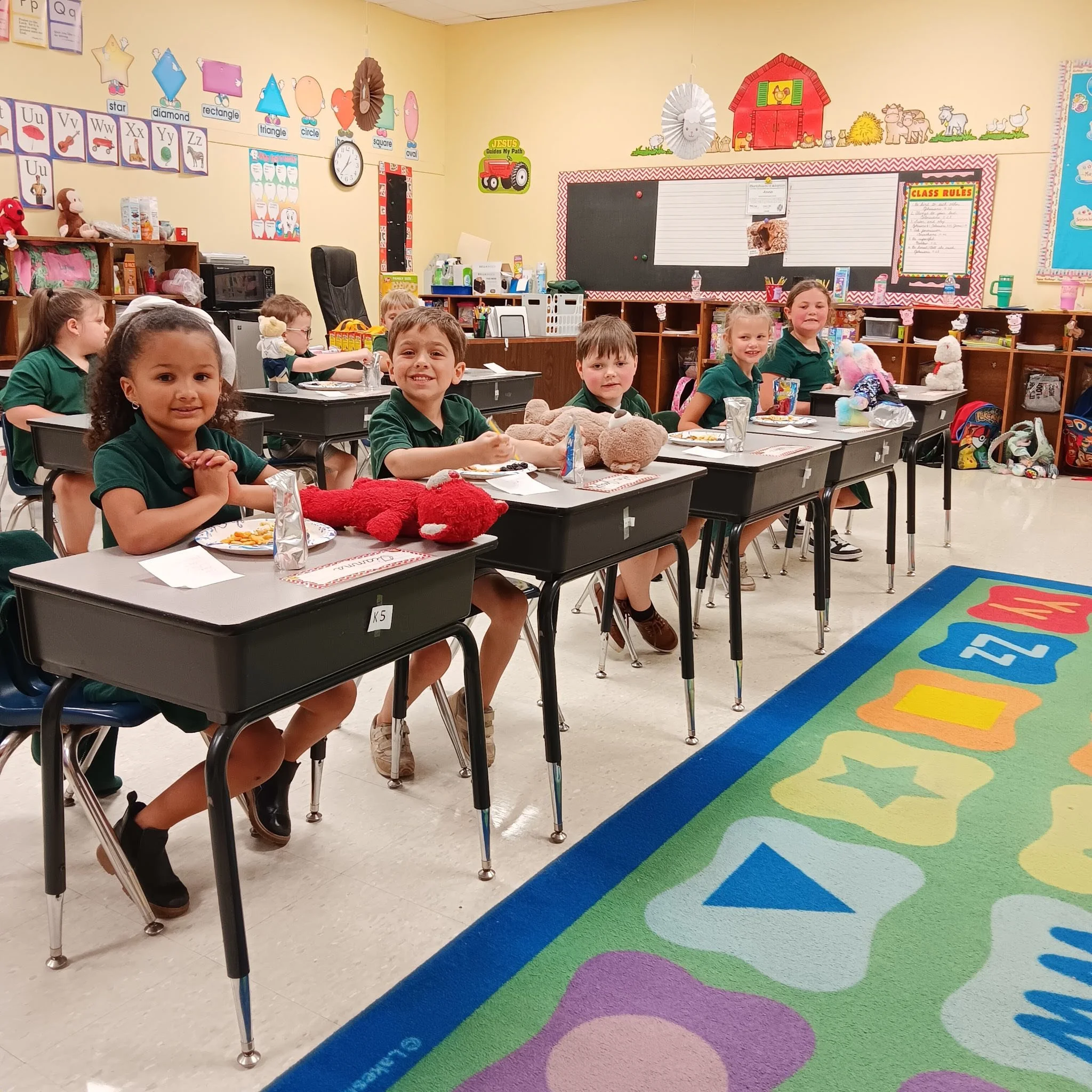 Young children sitting at desks in a classroom, smiling, with stuffed animals on their desks, colorful posters on the walls, and a bright, welcoming environment.