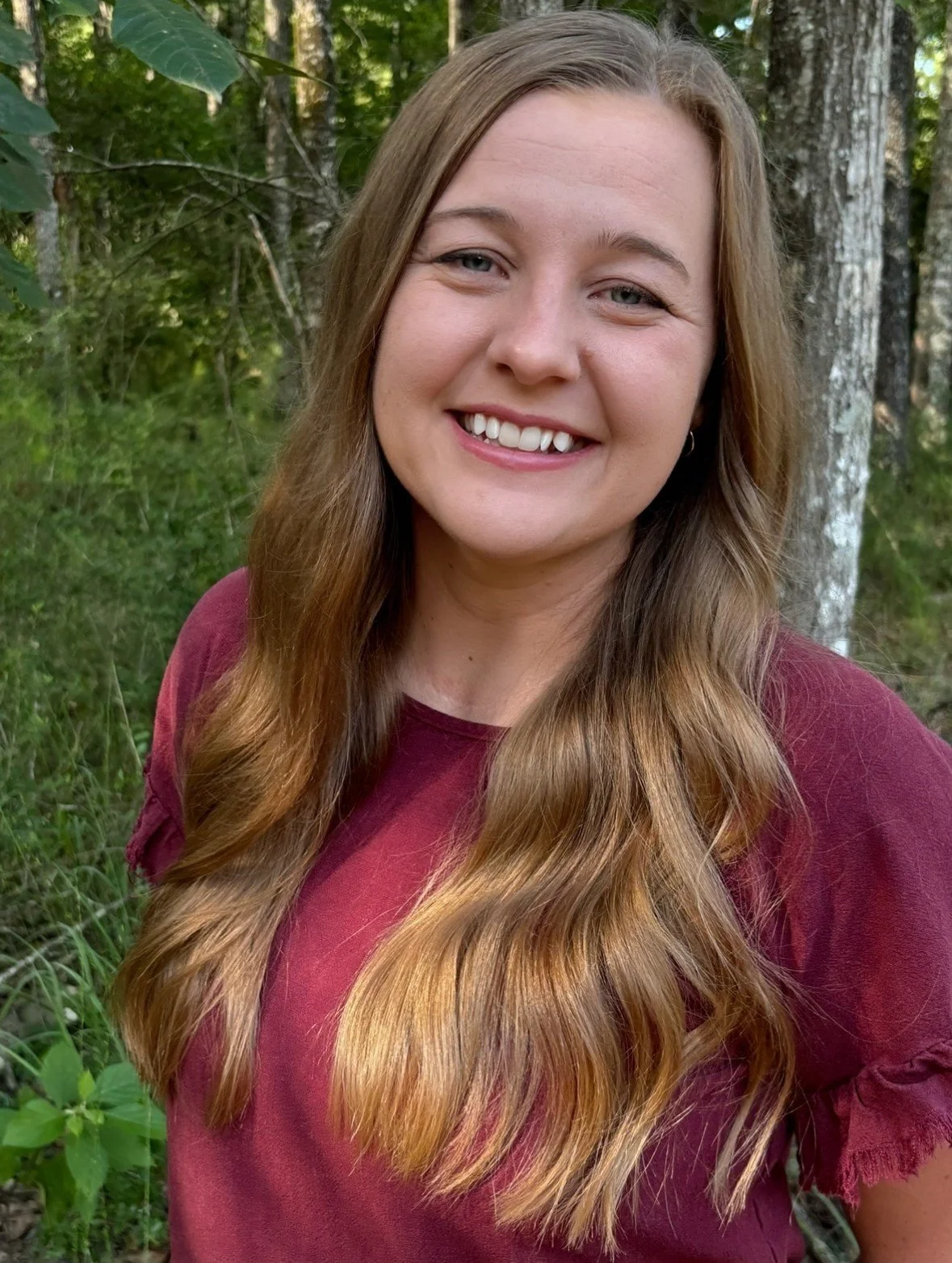 A woman with long wavy light brown hair and blue eyes smiling outdoors surrounded by green trees and foliage.
