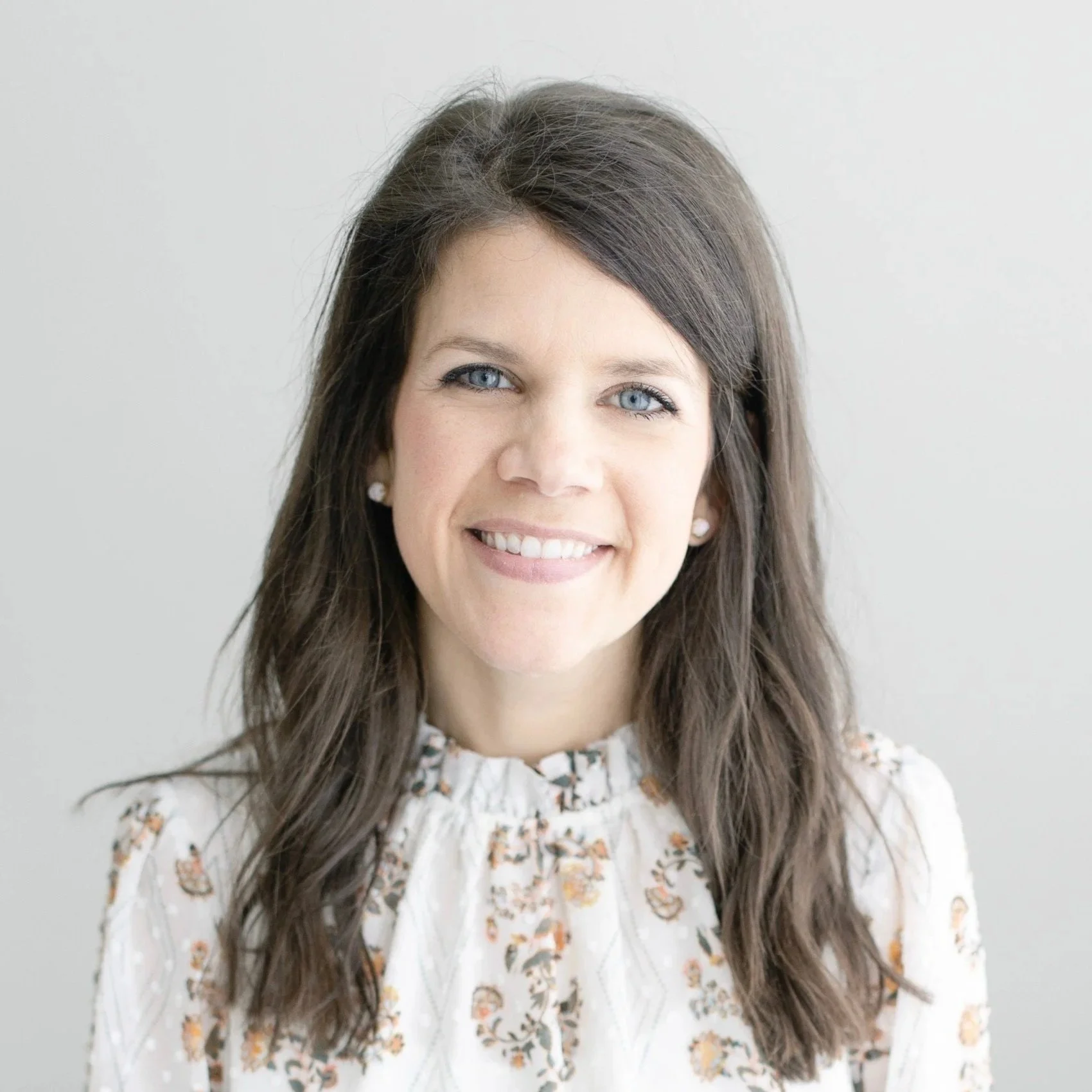 Portrait of a smiling woman with blue eyes, long brown hair, wearing pearl earrings and a floral patterned blouse, against a plain light background.