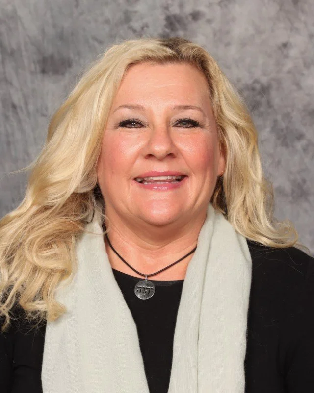 A woman with long blonde hair, smiling, wearing a black top, a light-colored scarf, and a black necklace with a round pendant, against a gray background.