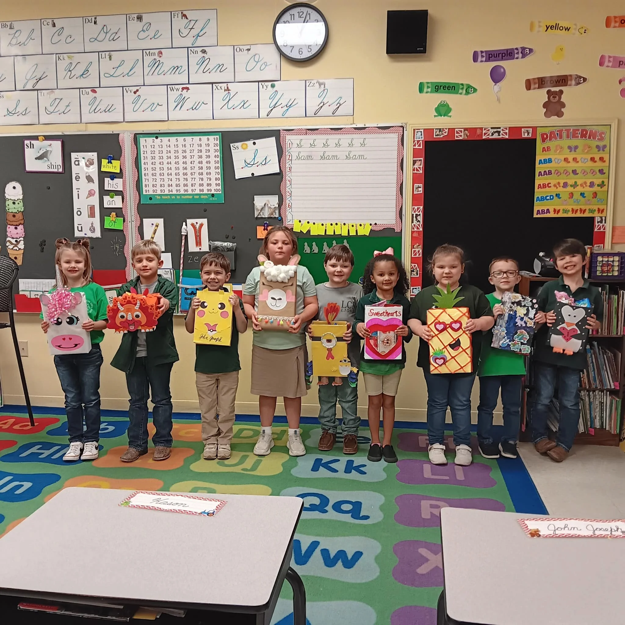A group of nine young children standing in a classroom, holding colorful valentine's day themed crafts. The children are smiling and standing in a row on a multicolored alphabet-themed rug, with educational posters, a clock, and classroom items visib
