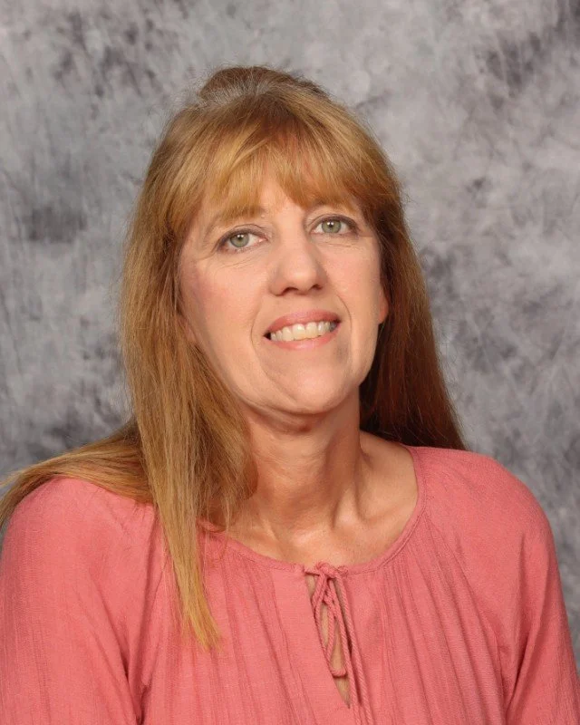 A woman with long red hair, light skin, and green eyes wearing a pink blouse, smiling at the camera against a gray textured background.