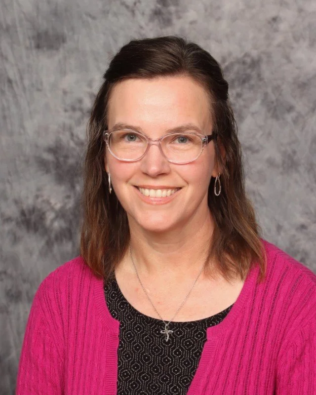 Smiling woman with shoulder-length black hair wearing a blue top, standing in front of green plants indoors.