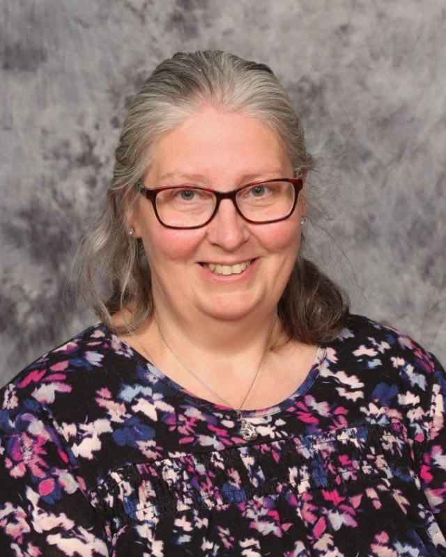 A woman with shoulder-length black hair smiling in a greenhouse or indoor garden with lush green plants in the background.