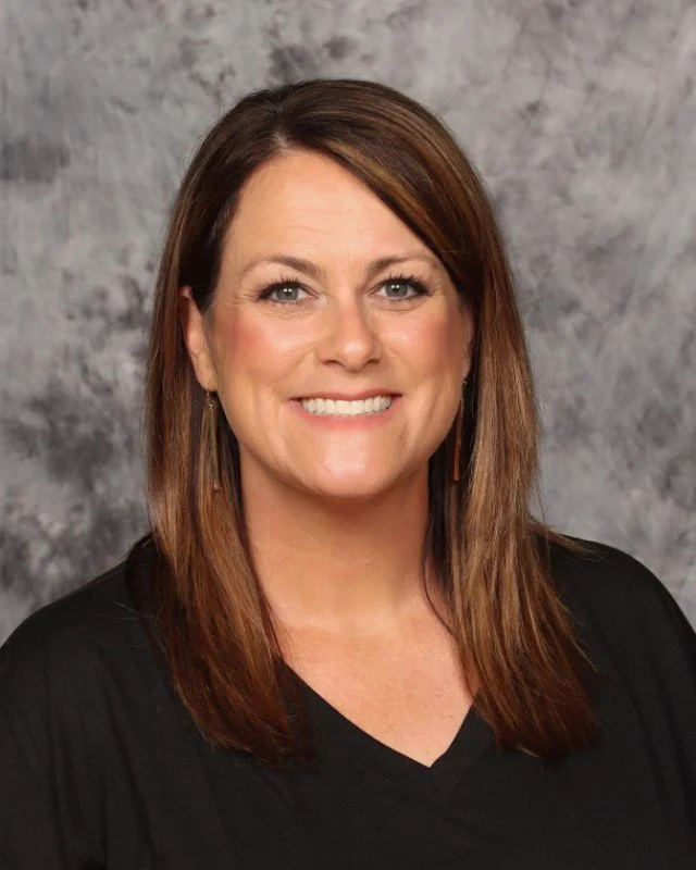 A woman with shoulder-length brown hair and blue eyes smiling, wearing a black top, in front of a gray textured background.
