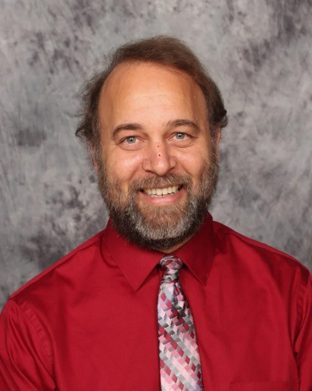 A man with dark curly hair, glasses, and a beard looking slightly to the right, wearing a dark green sweater, in a bright room with a potted plant in the background.