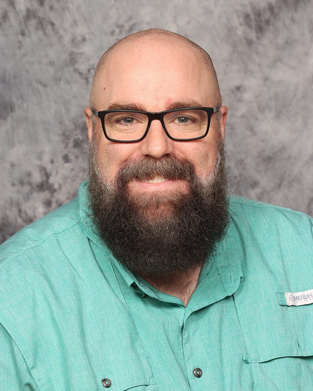 A middle-aged man with glasses, a full beard, and a mustache, smiling, wearing a teal button-up shirt with a name tag, against a gray mottled background.