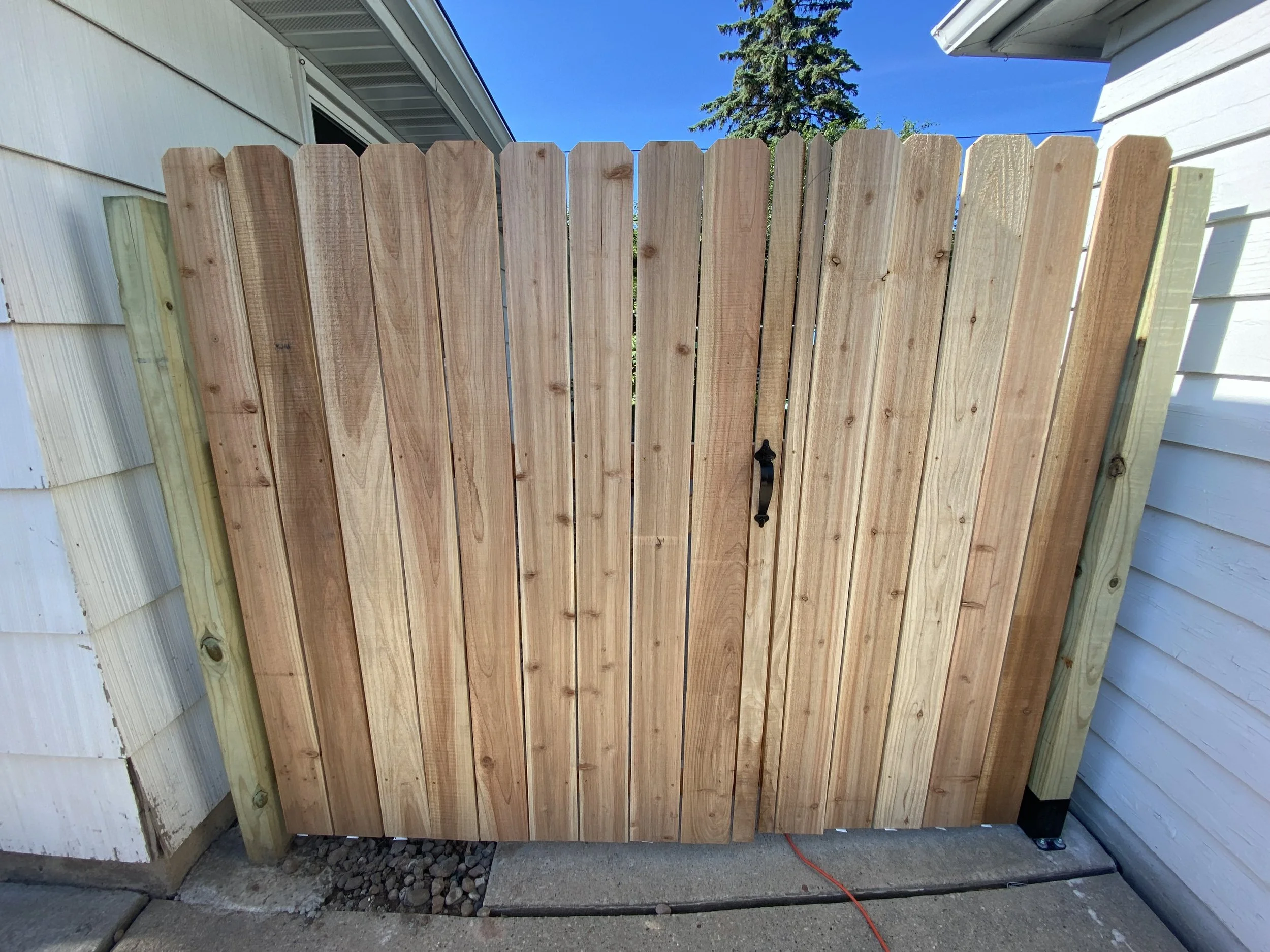 New wooden backyard gate installed between two white houses, with a black handle and a latch, under a clear blue sky.