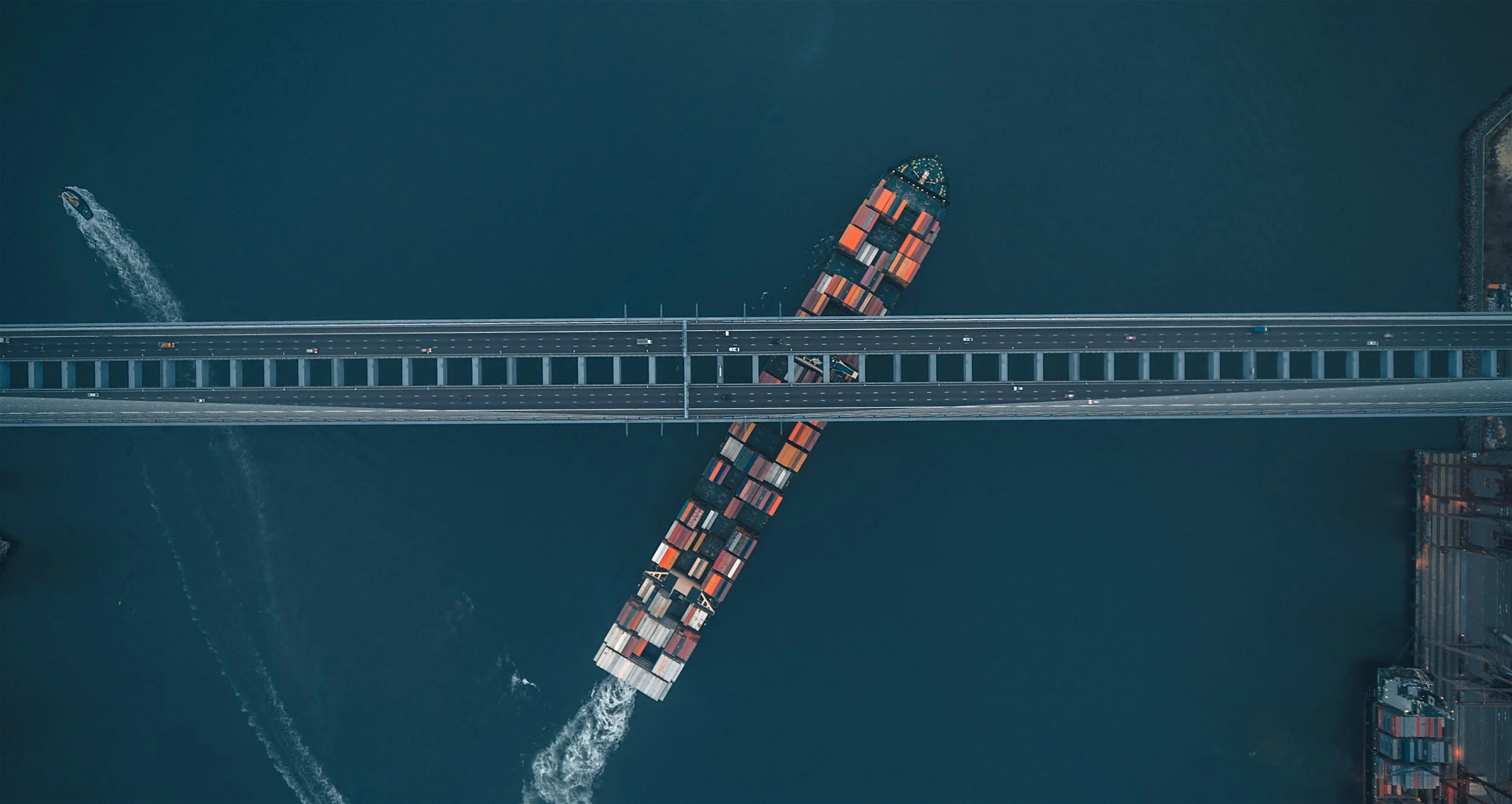 An aerial view of a cargo ship carrying containers passing underneath a long bridge on a body of water.