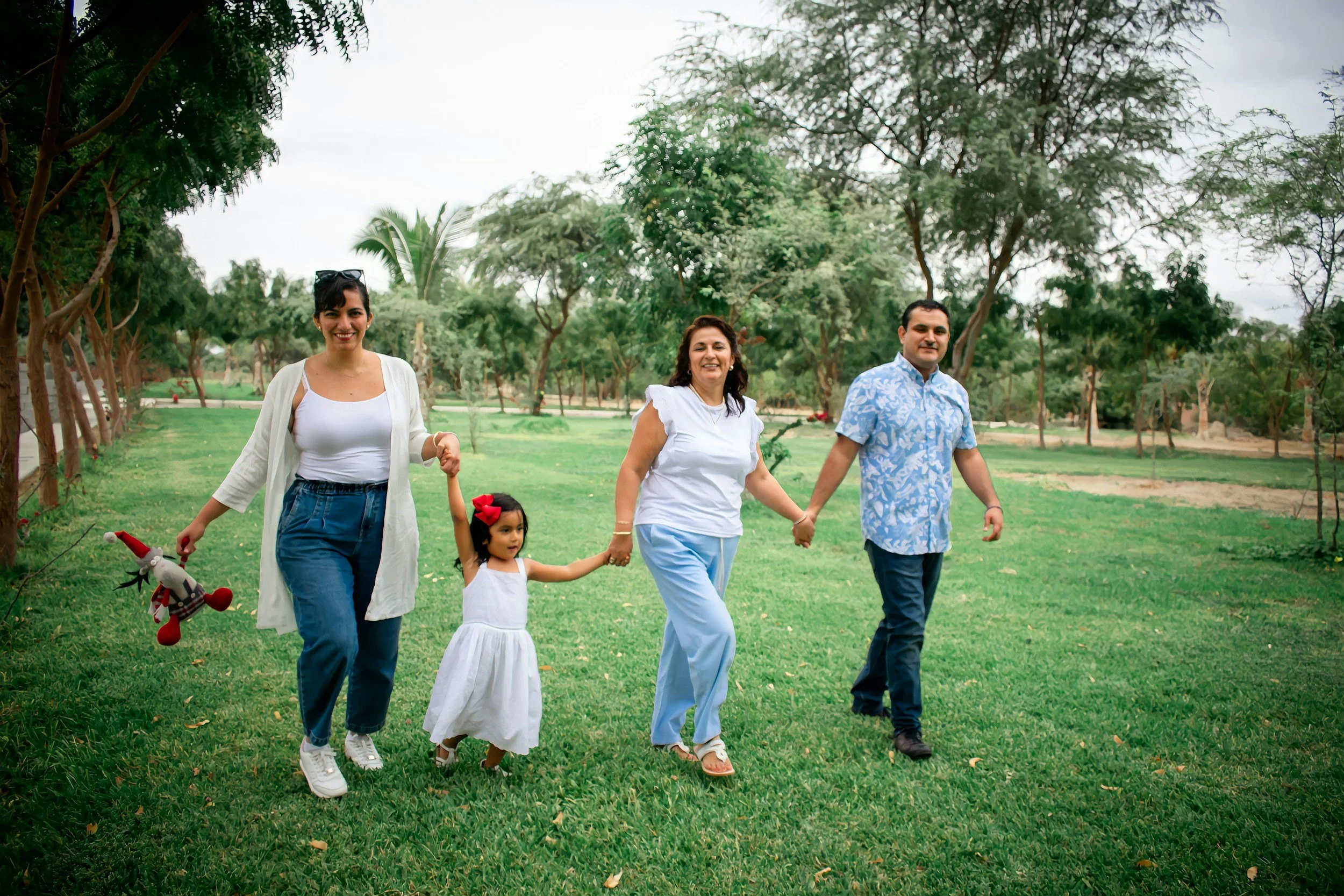 Family of four holding hands and walking in a park with green grass and trees, smiling and enjoying a casual day outdoors.
