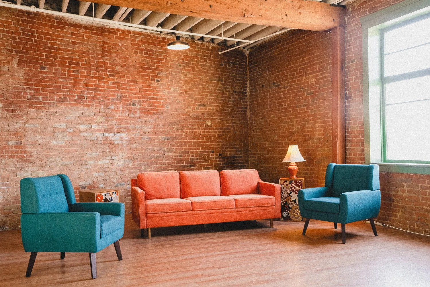 Orange sofa and teal chairs beside exposed brick and a sunlit window