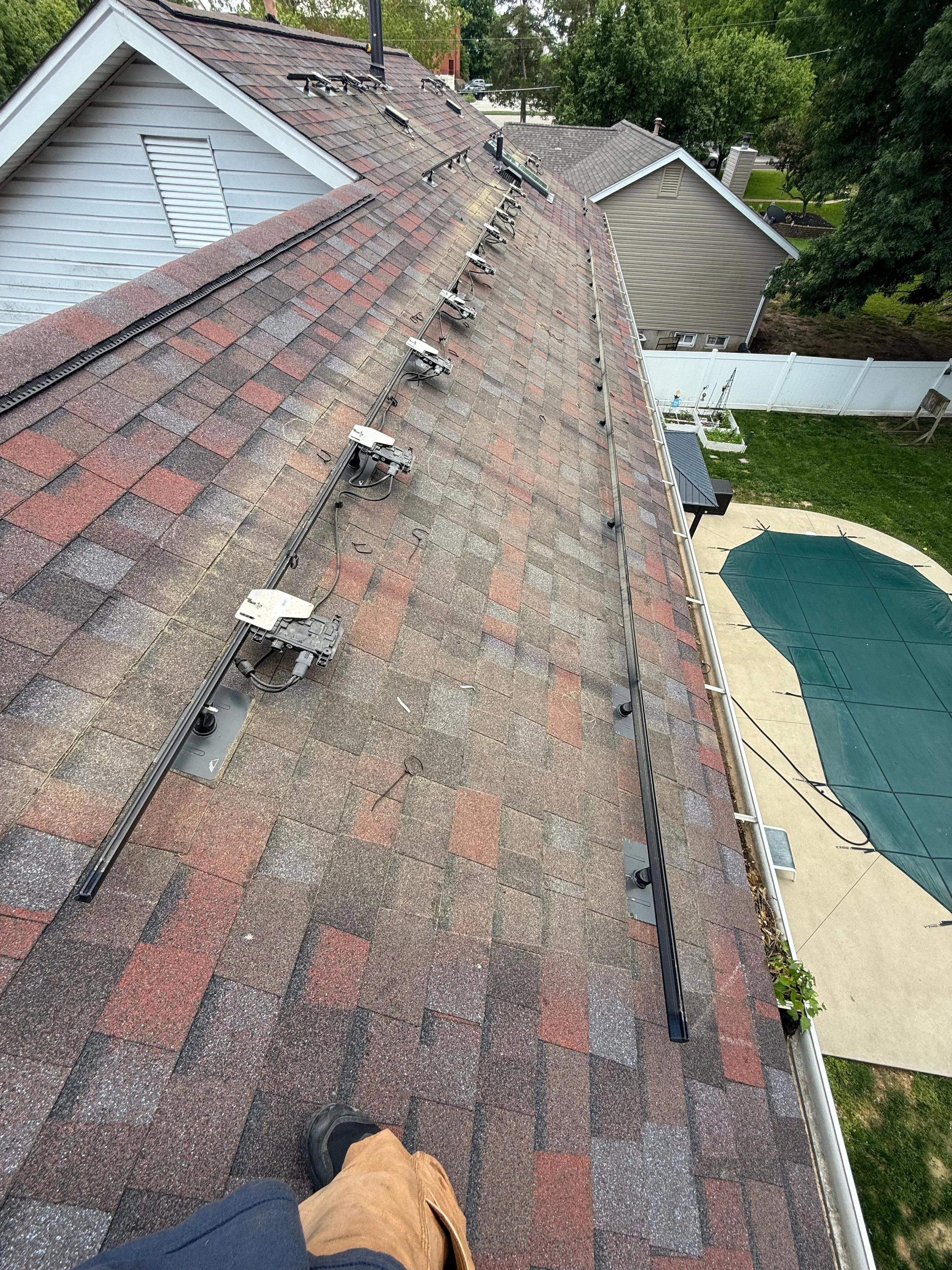 View of a residential roof with mounted solar panel equipment and wiring, overlooking the backyard with a covered patio and white fence.