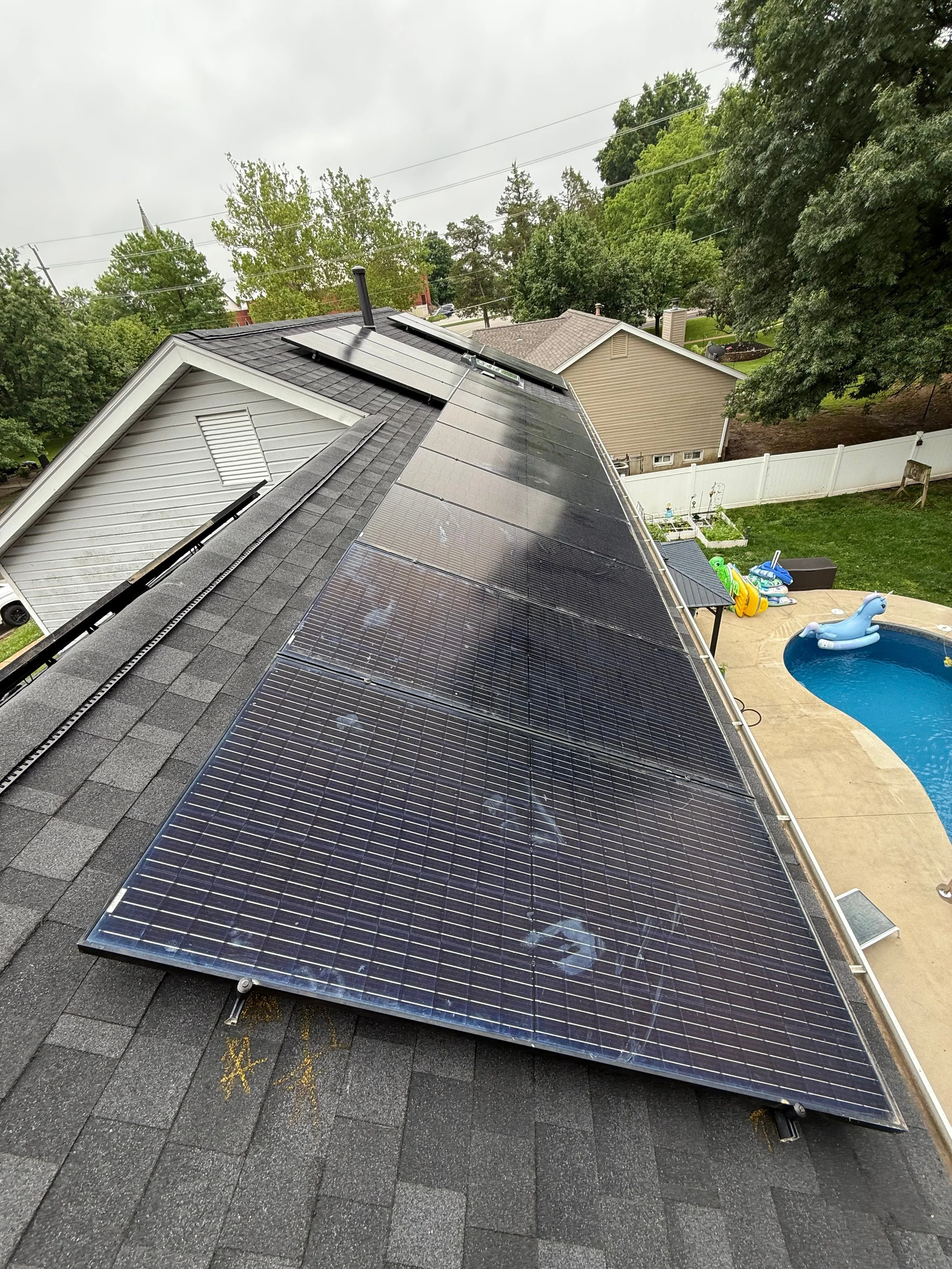 Residential roof with solar panels, neighboring houses, backyard pool with pool toys, overcast sky.