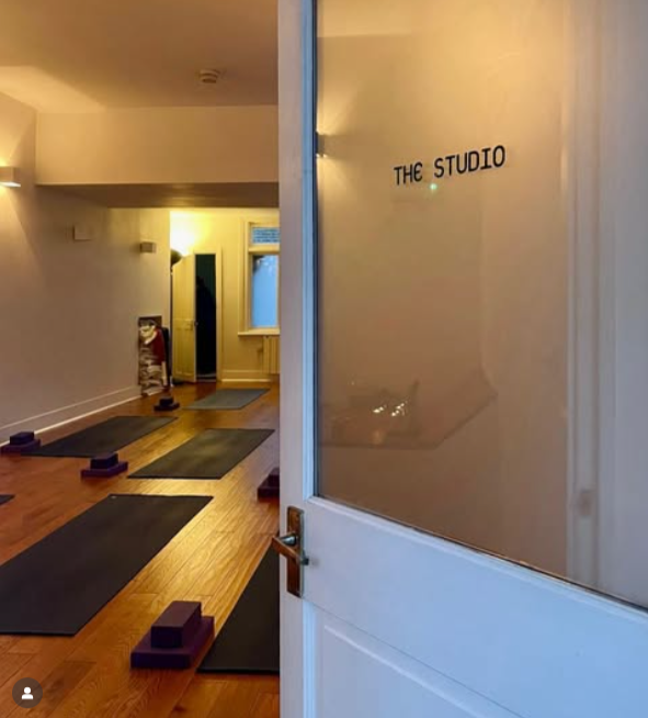 View of a yoga or exercise studio with black mats and purple blocks arranged on a wooden floor, seen through a partially open door with 'THE STUDIO' written on the glass.
