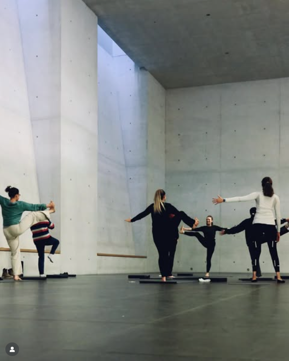 A group of people practicing yoga in a studio with tall concrete walls. Some are stretching while others hold hands in a circle.