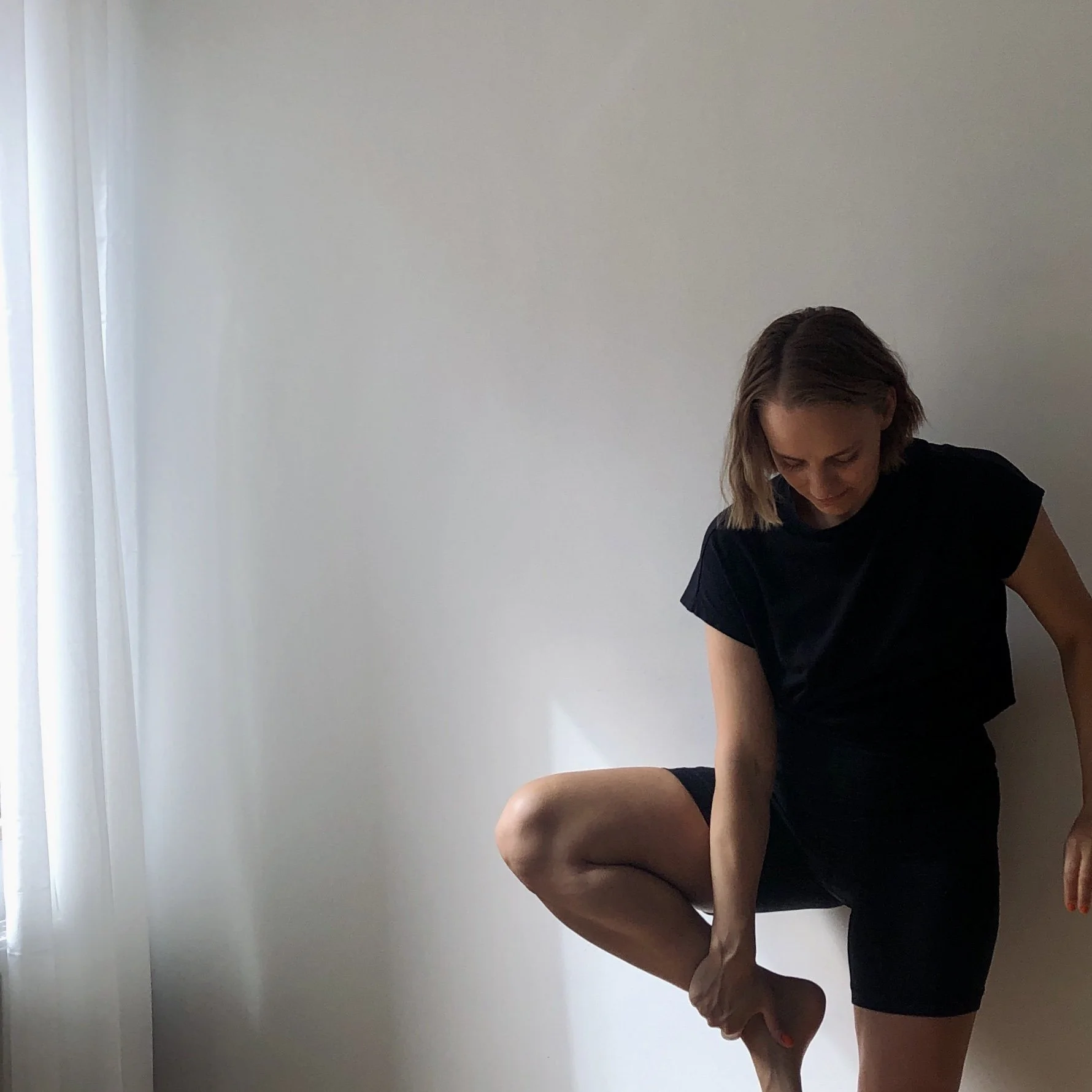 Woman in black shirt and shorts standing against a plain white wall, holding her ankle in a stretching pose.