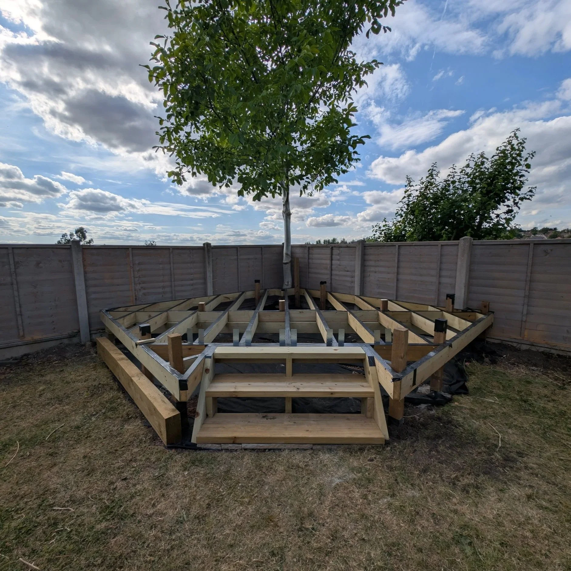 A backyard with a partially built wooden deck around a tree, with steps leading up to the deck, enclosed by a wooden fence under a partly cloudy sky.