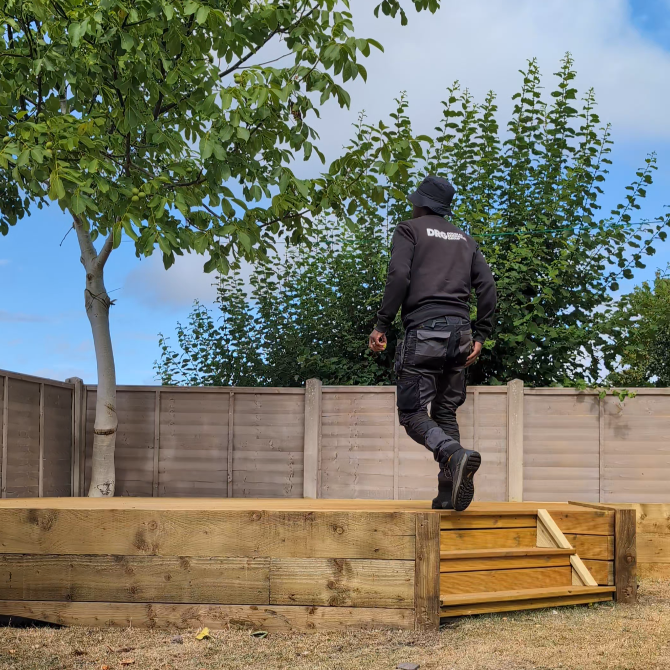 DRG contractor dressed in black work clothing with a logo on the back is walking up a small wooden staircase on a raised garden bed in a backyard with a fence, trees, and a cloudy sky.