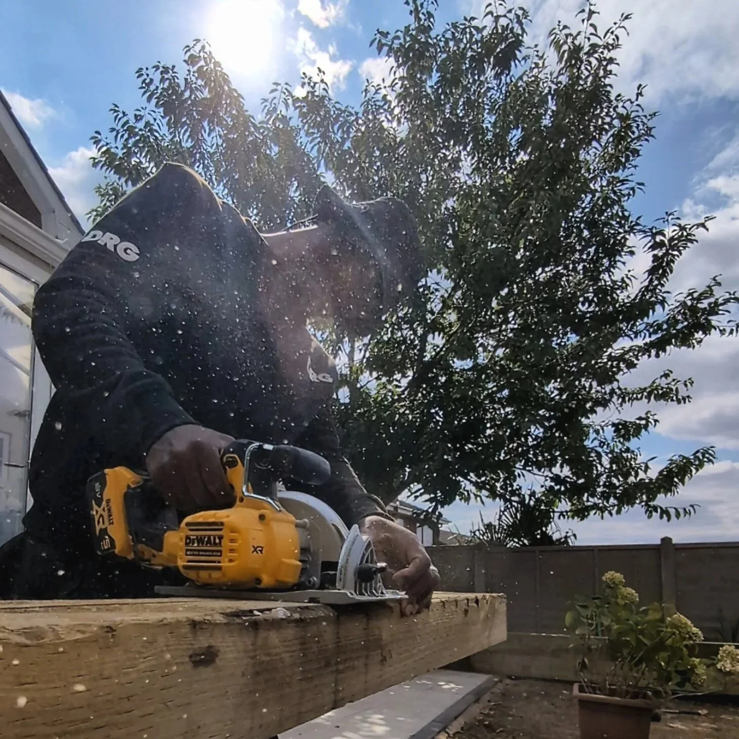 A DRG contractor using a yellow DEWALT cordless circular saw to cut a piece of wood outdoors, with sunlight and a tree in the background.