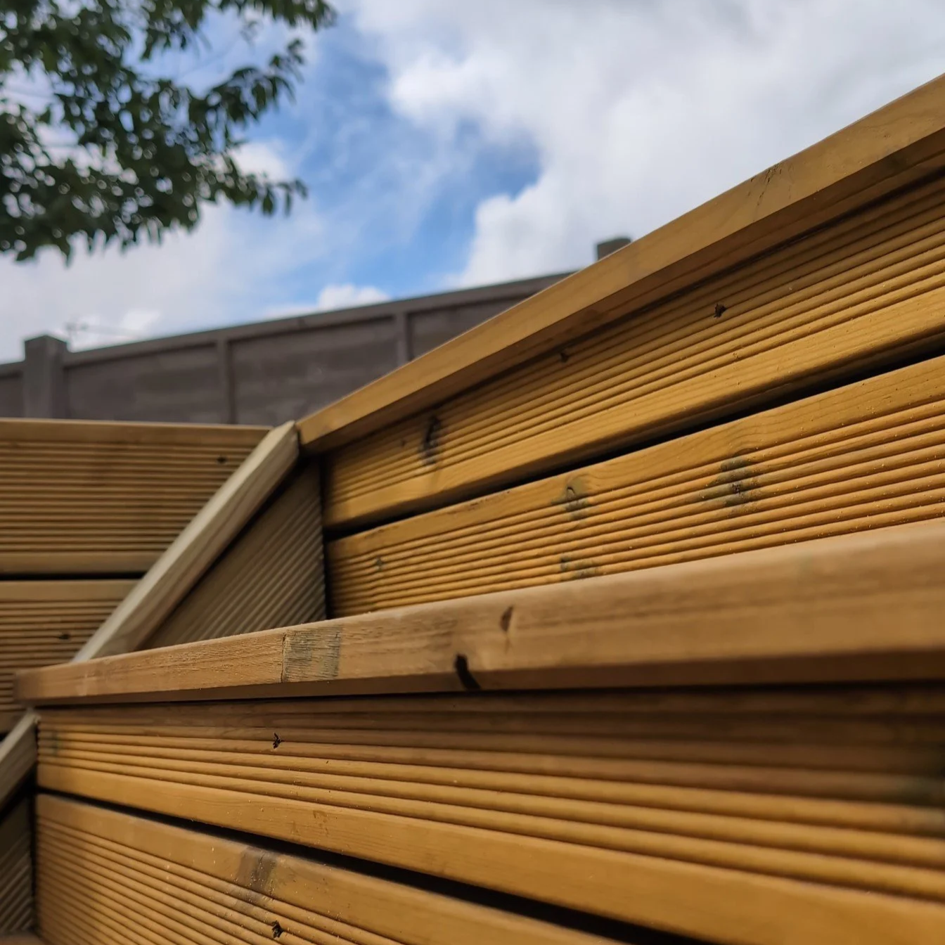 Close-up of wooden decking detail with a fence, a tree, and blue sky with a few clouds in the background.