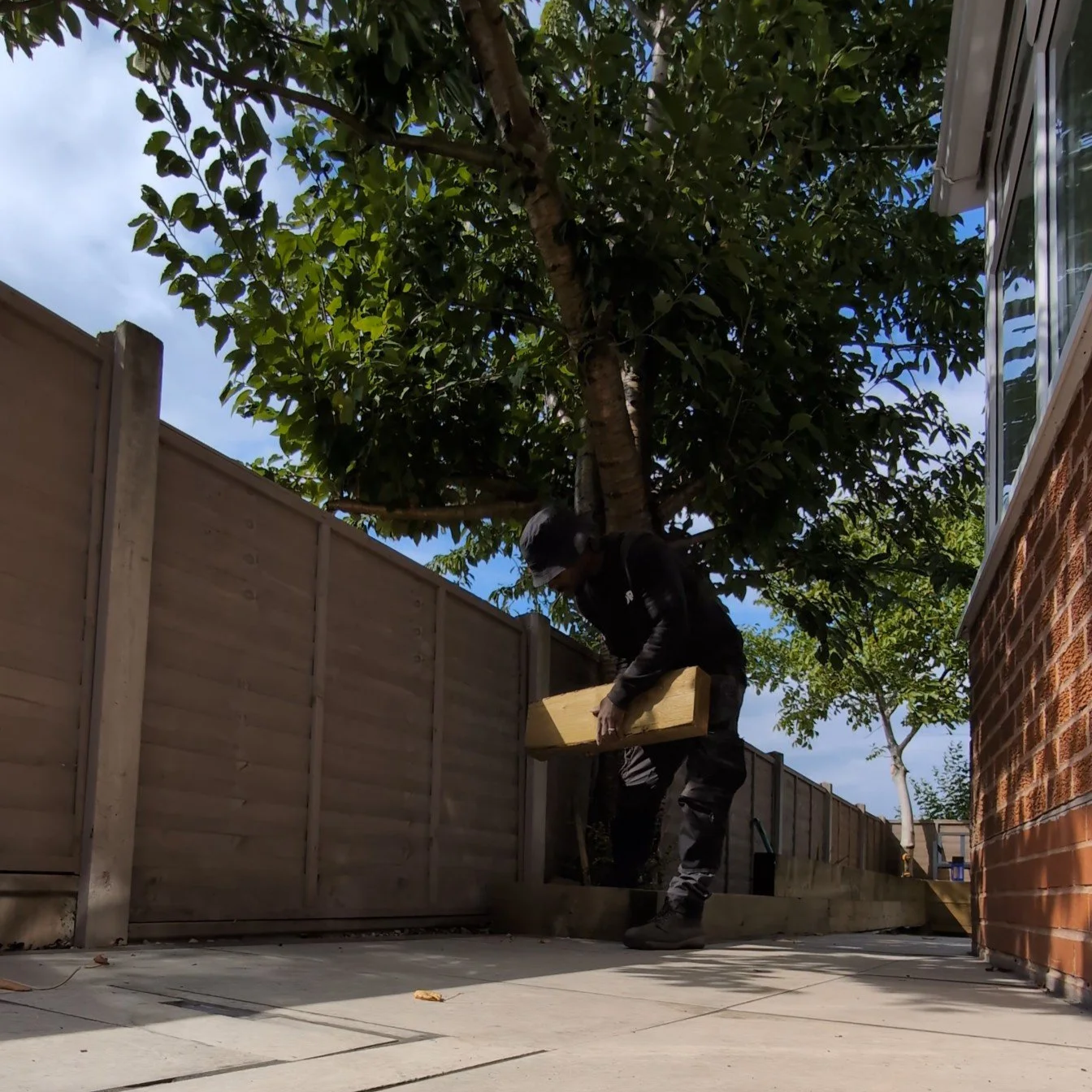 DRG contractor working on a raised garden bed or platform outdoors next to a wooden fence, with trees and a brick house wall nearby.