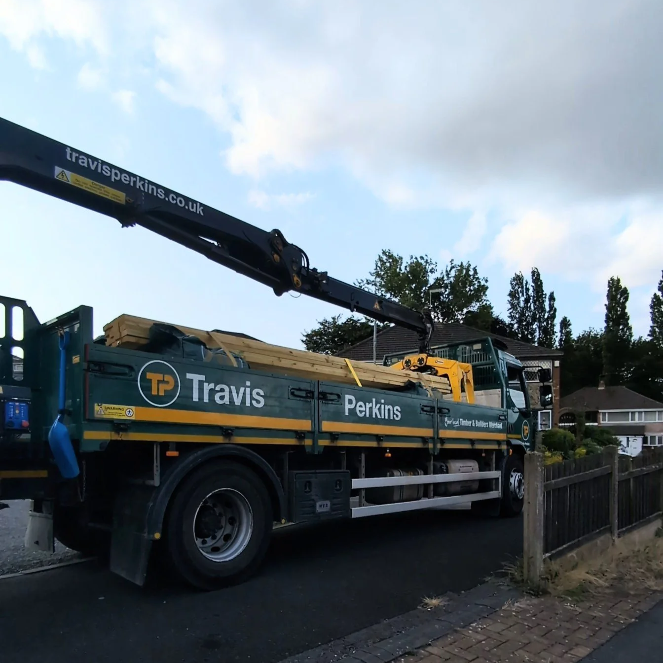A green flatbed truck with the branding 'Travis Perkins' and a crane arm extended over the truck bed, carrying wooden planks, parked on a residential street.