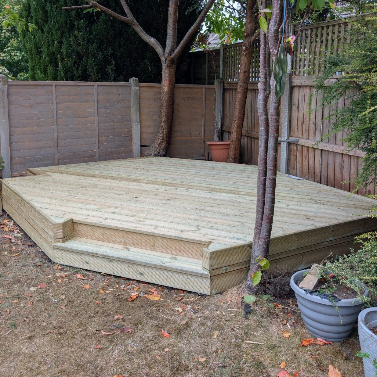 A newly built wooden deck in a backyard, surrounded by a wooden fence, with trees and potted plants nearby.
