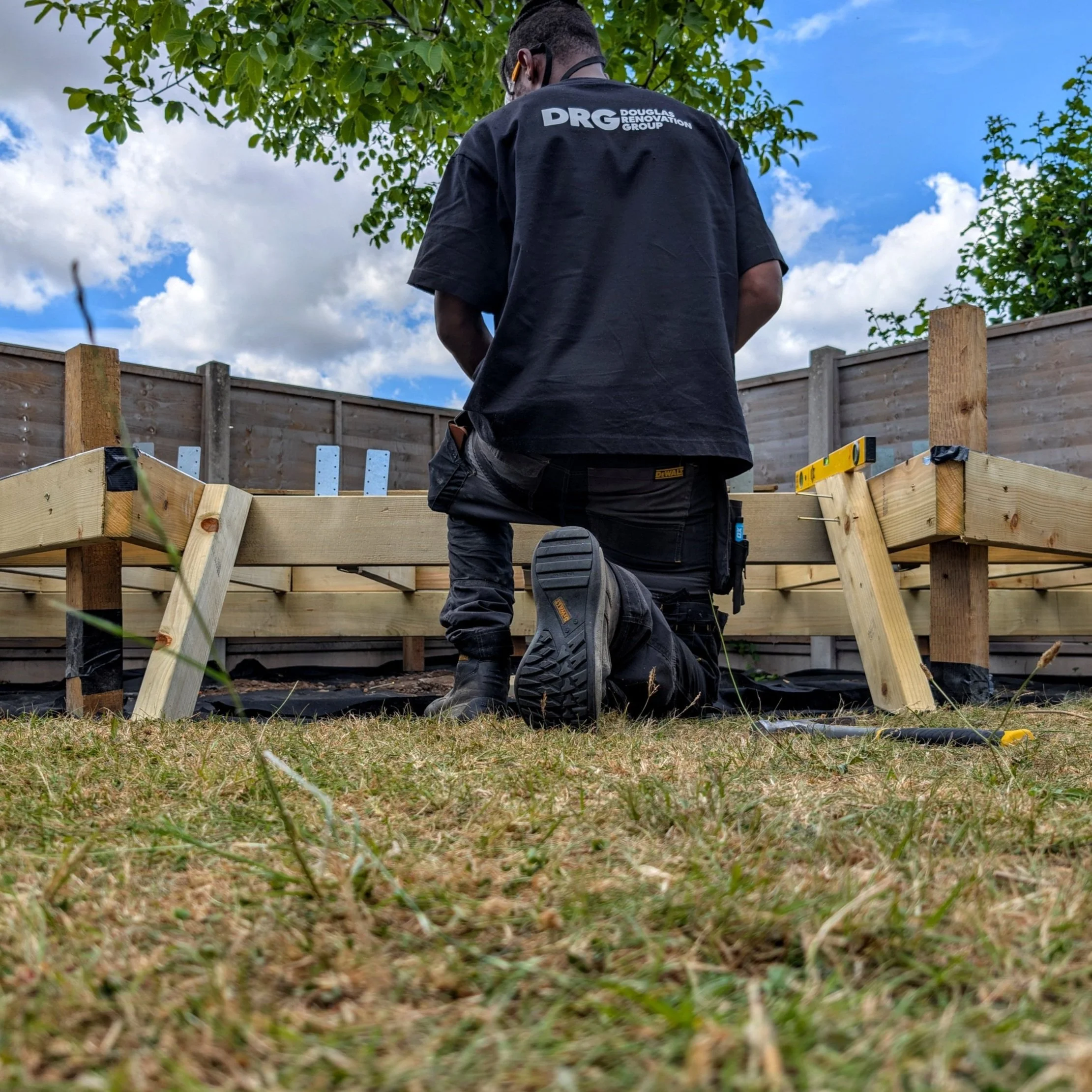 DRG contractor kneeling on the ground, working on a wooden deck structure outdoors under a blue sky with scattered clouds.
