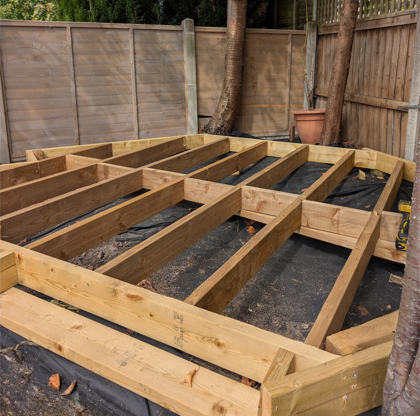 Wooden framework for a deck construction in a backyard, surrounded by a wooden fence and trees.