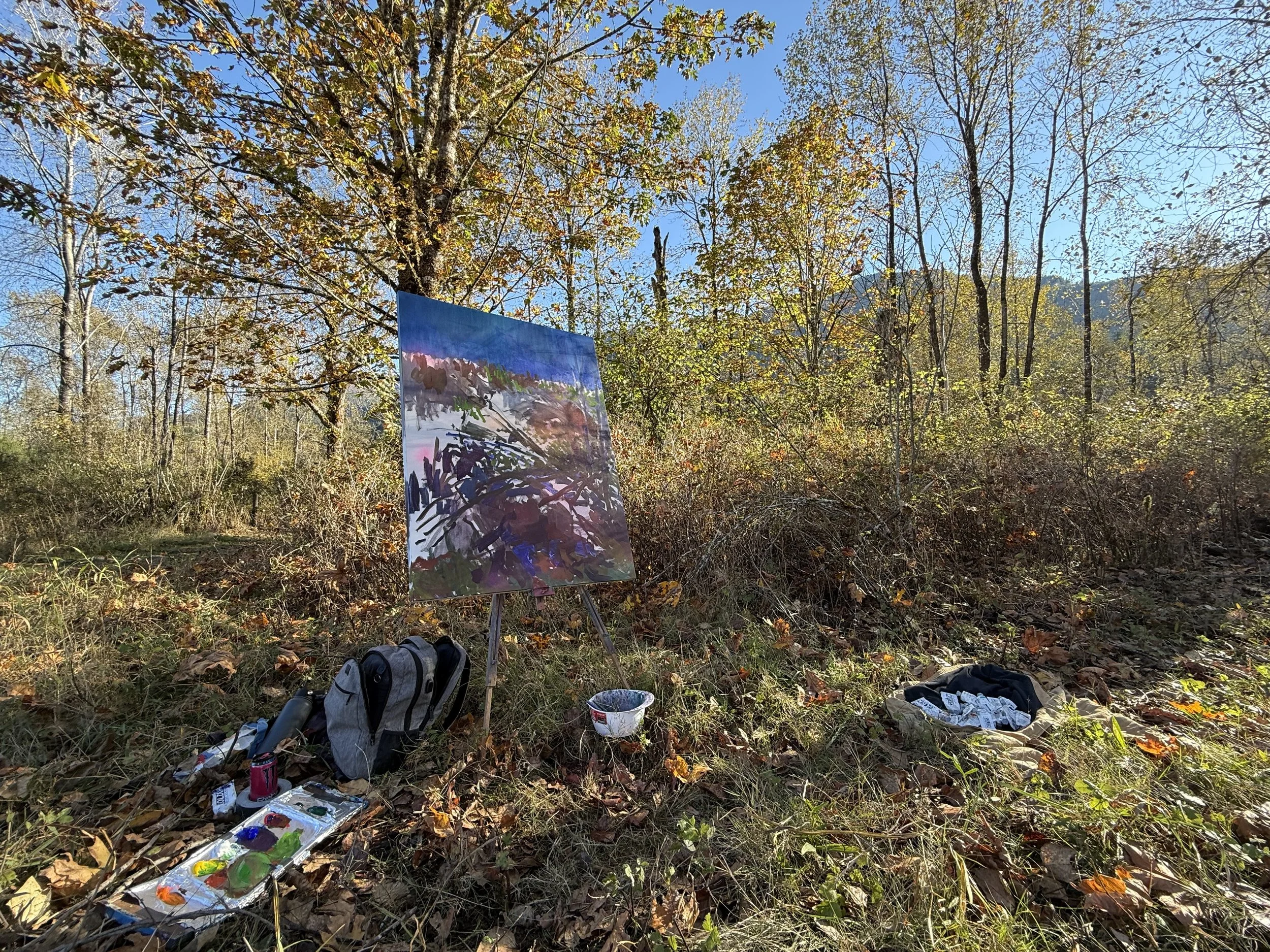 An outdoor scene in a wooded area with a painting on an easel, various art supplies, and a backpack on the ground. The trees have autumn leaves, and the sky is blue.