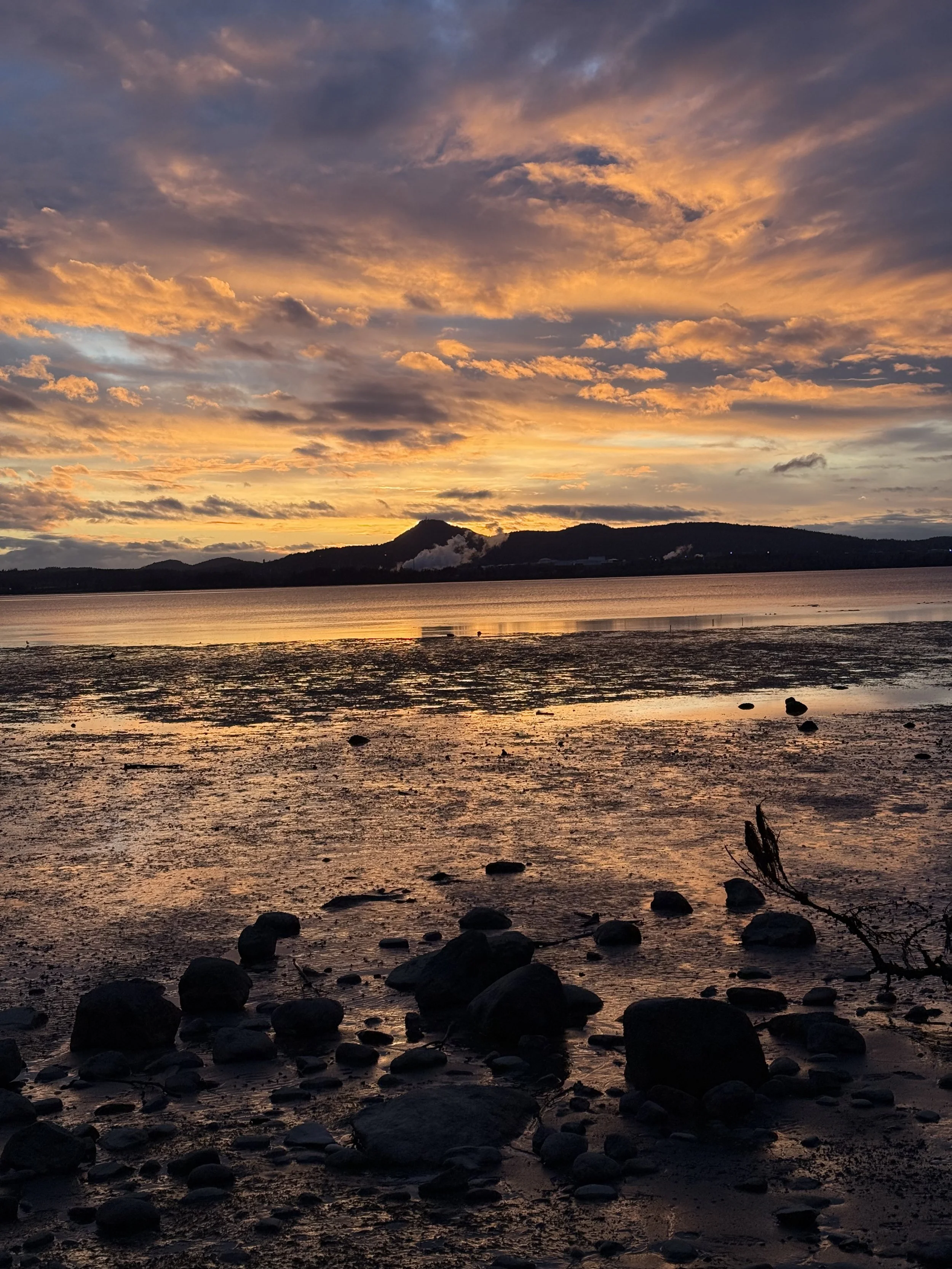 Sunset over a calm body of water with a rocky shoreline and distant hills, colorful clouds in the sky looking across at Anacortes, Washington State