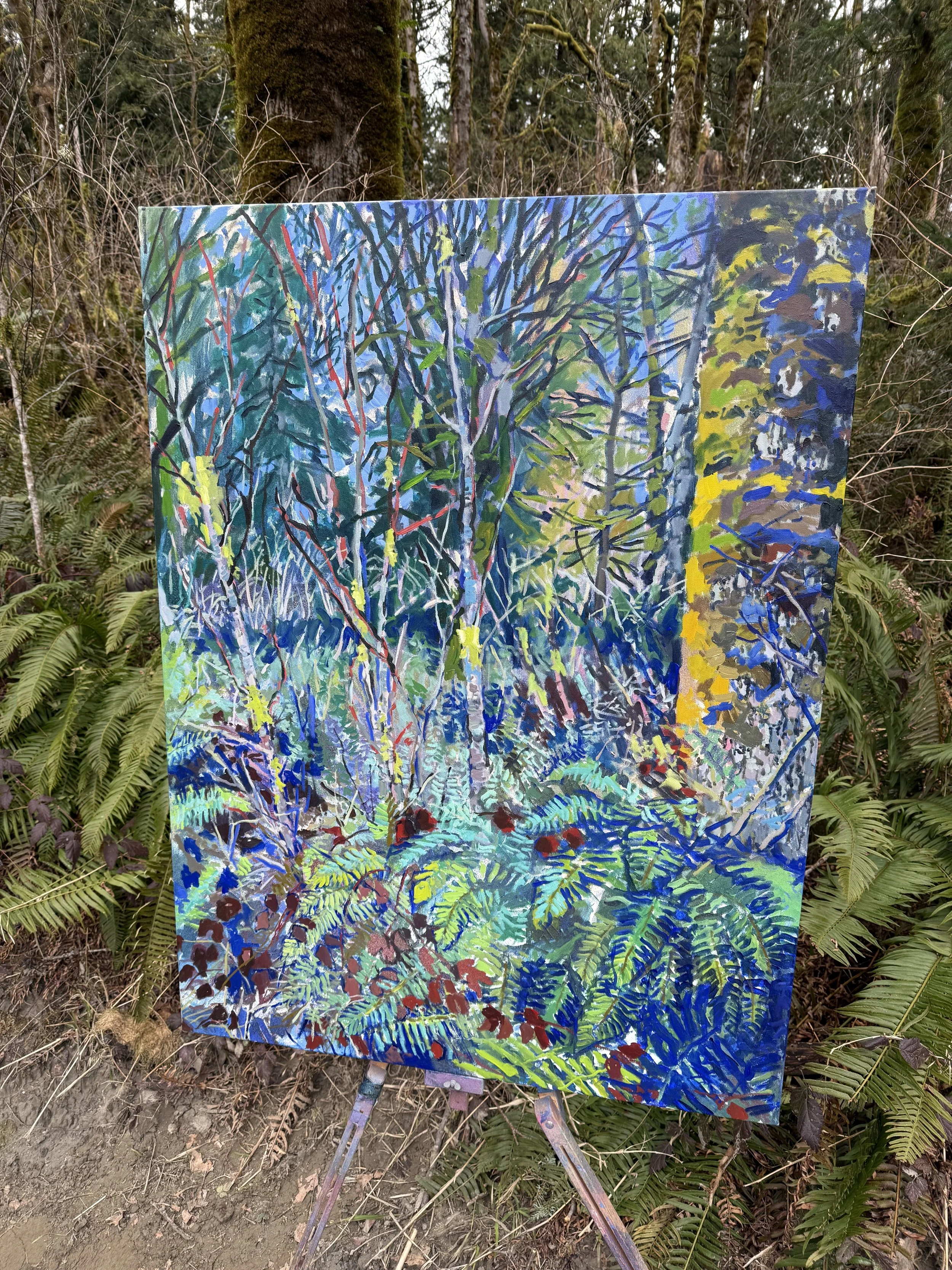 A colorful large scale plein air painting of a forest scene with trees, bushes, and ferns, displayed on an easel outdoors in a wooded area located in Razar State Park