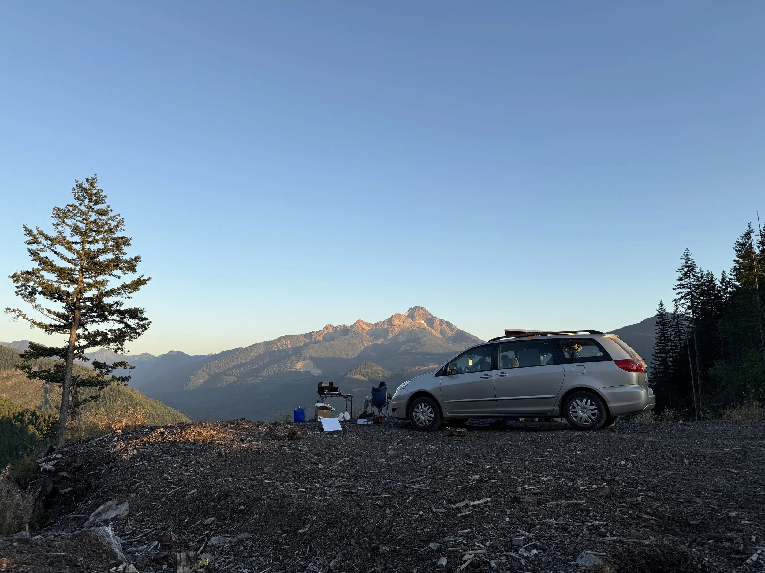 A silver minivan parked on dirt with a scenic mountain view in the background, including a tree on the left and a clear blue sky.