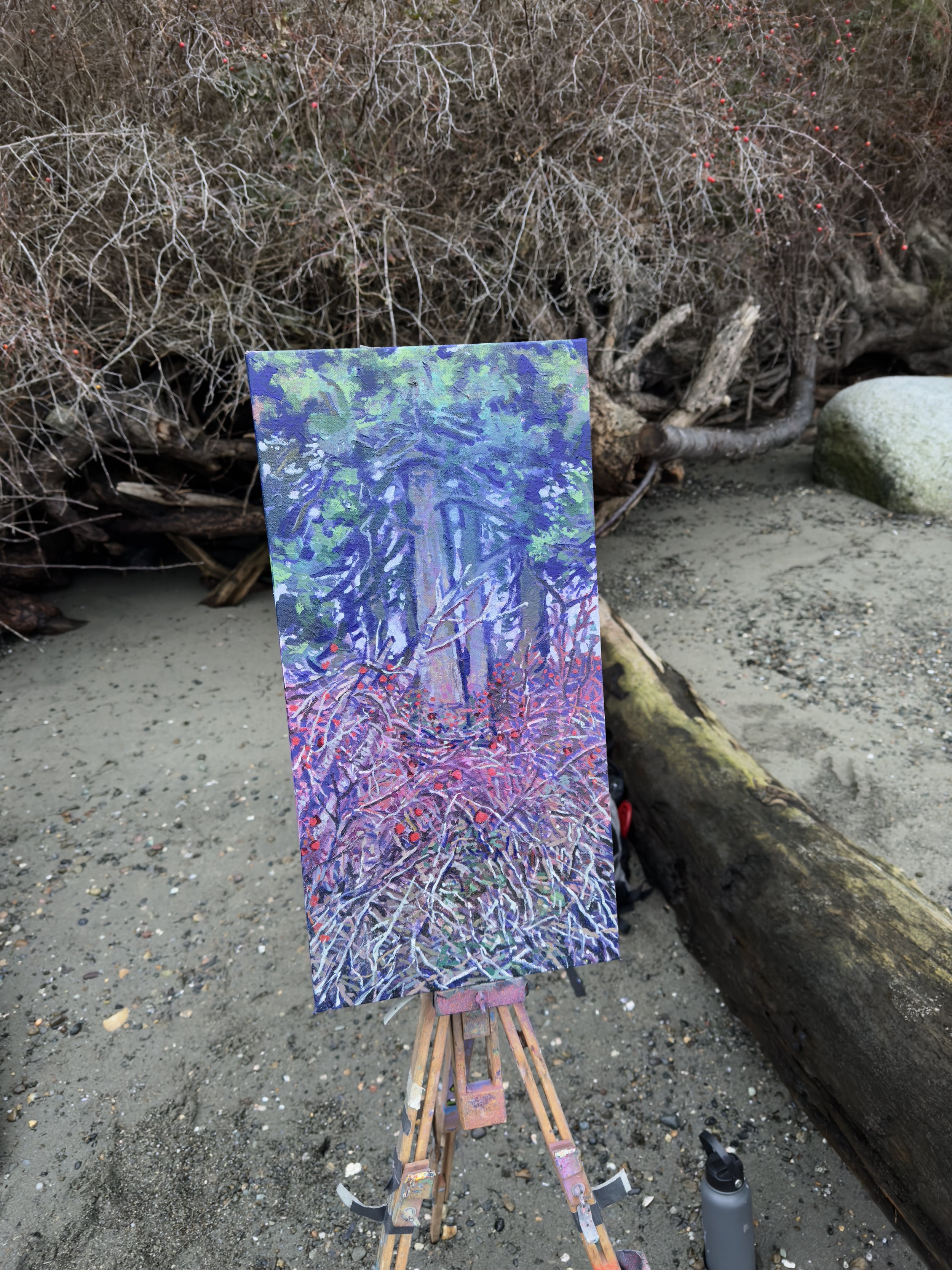 A colorful painting of a forest scene on a canvas, set on an easel on a beach with sand, rocks, driftwood, and leafless bushes in the background.