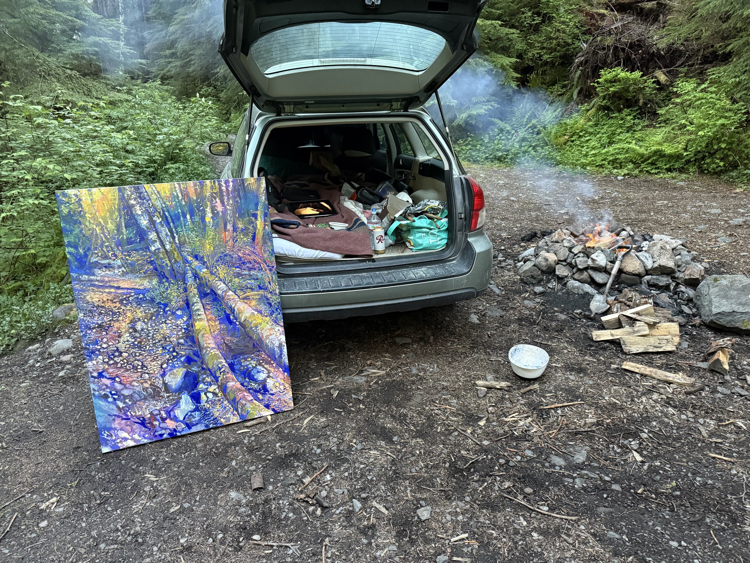 Open car trunk at a camping site in the woods, with art supplies and gear inside, next to a small campfire with rocks and a axe, and a colorful large scale plein air abstract painting leaning against the car.