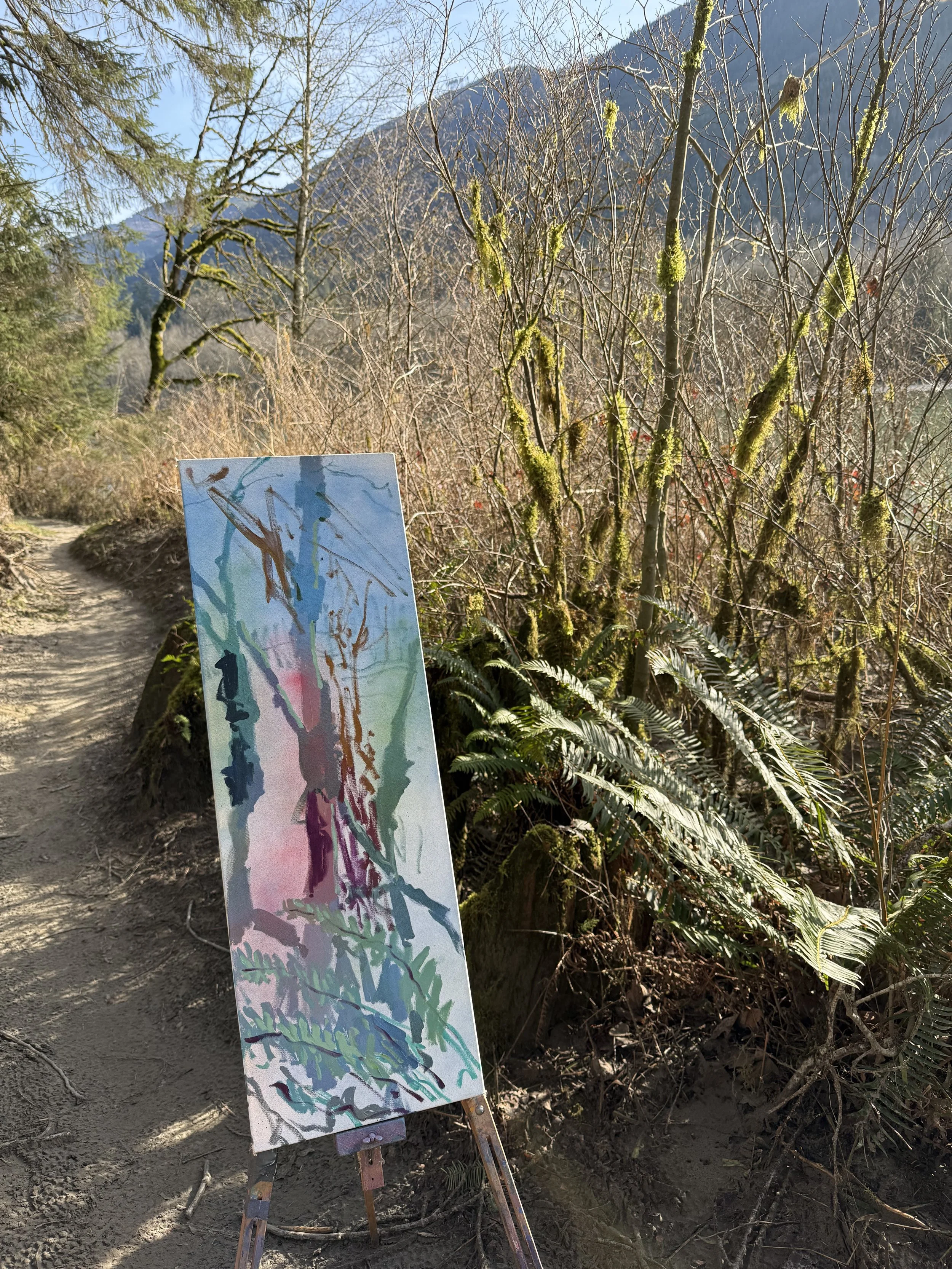 An abstract watercolor painting on an easel placed on a dirt trail in a forest with ferns, moss-covered branches, and mountains in the background.