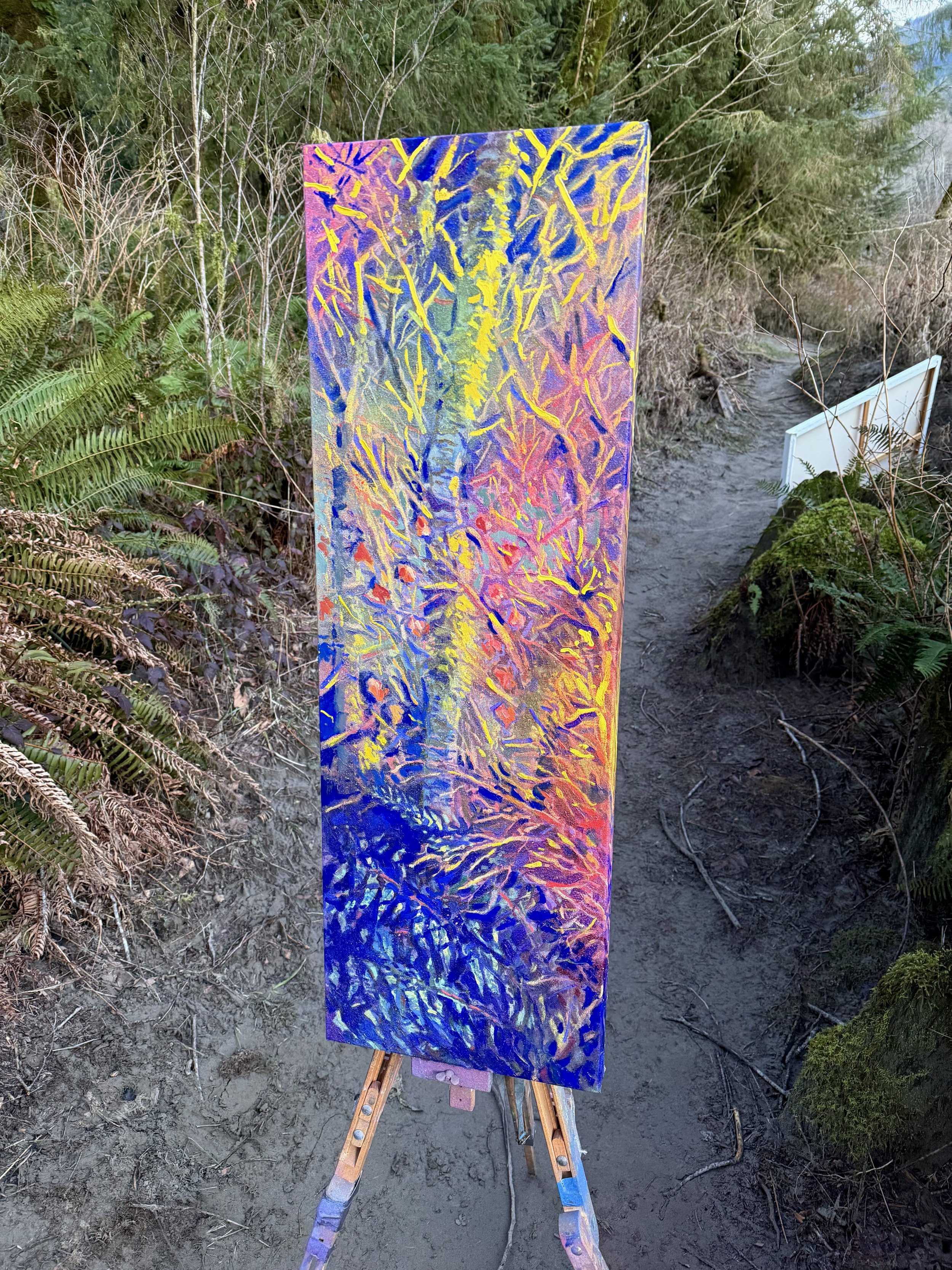 A colorful abstract painting on an easel outdoors in a forested area. The painting features vibrant yellow, pink, blue, and purple brushstrokes creating a textured, tree-like pattern.