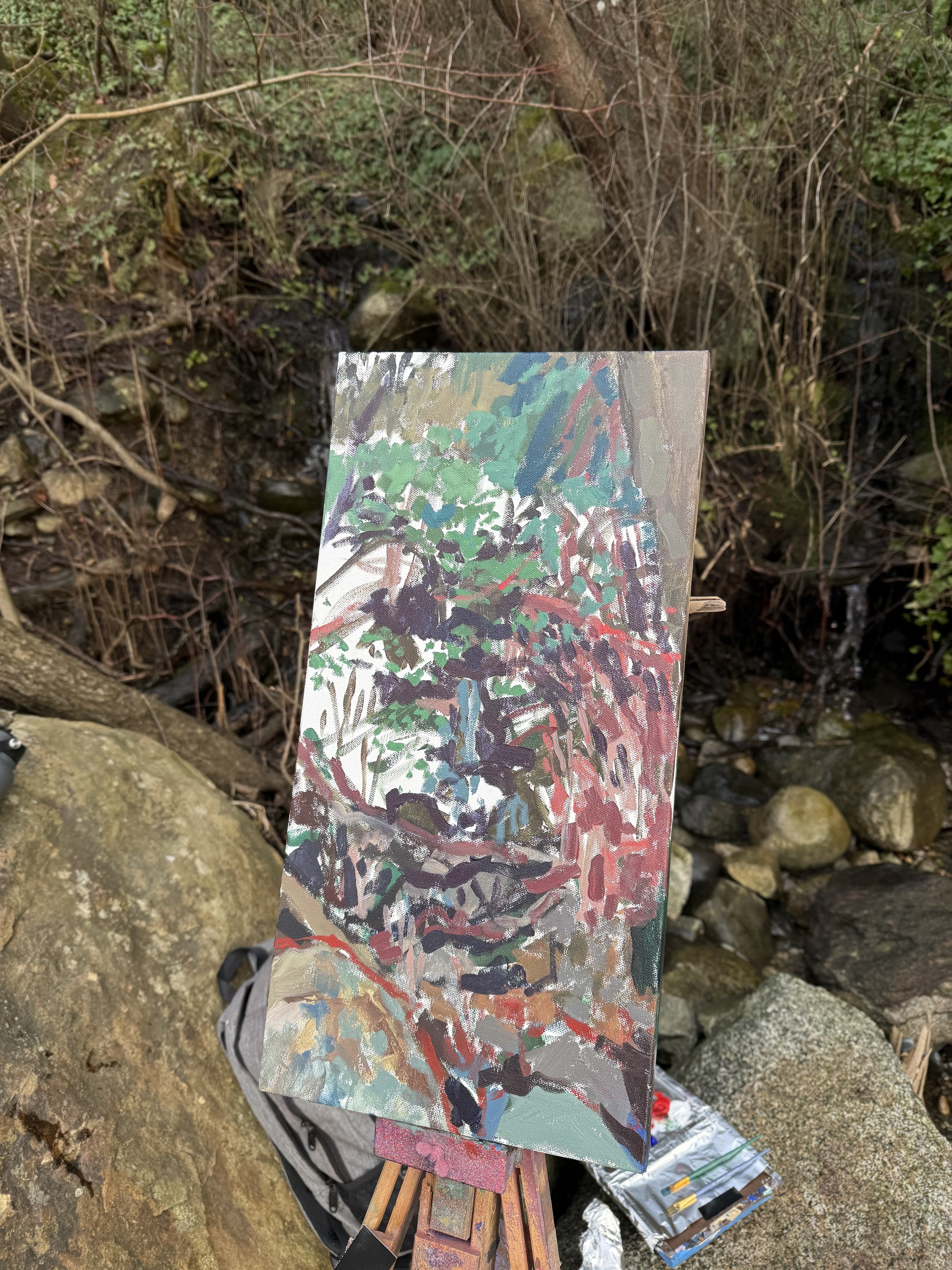 Painting of a forest scene with trees, rocks, and green foliage on an easel outdoors among rocks and vegetation.