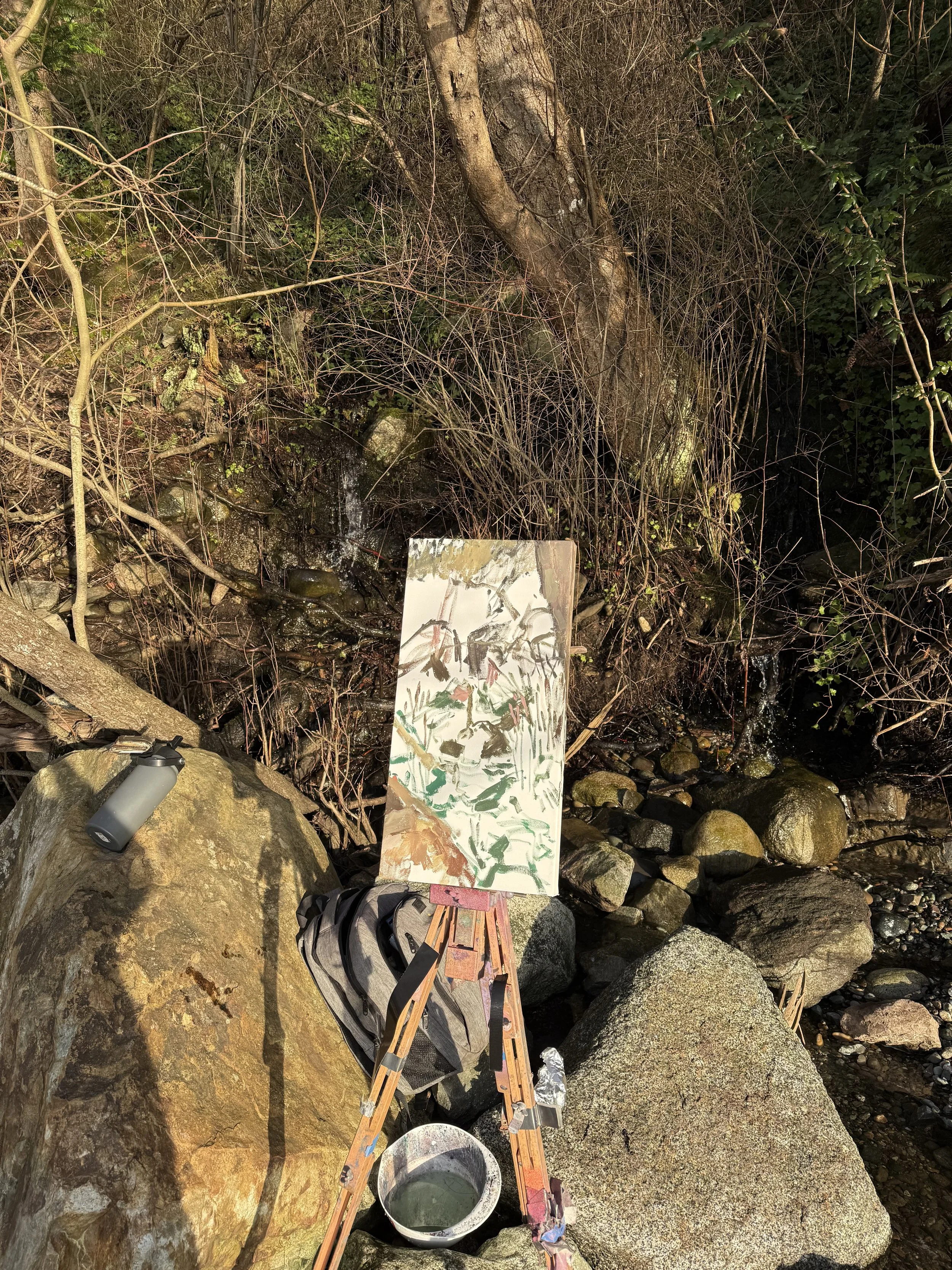 An outdoor painting setup with an easel, a partly painted canvas, a backpack, a water bottle on a large rock, a bucket of water, and rocks and trees in the background.