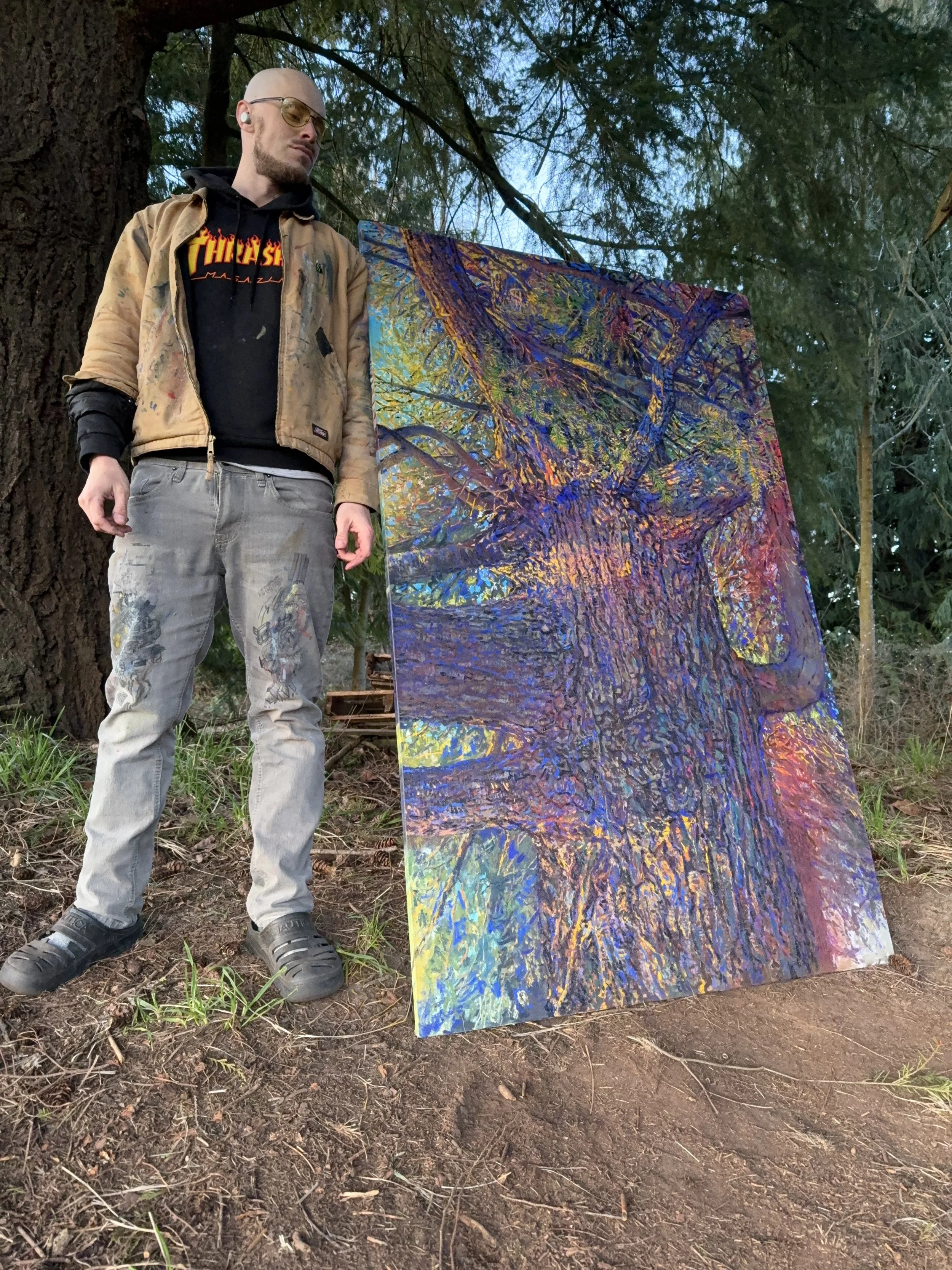 A man with sunglasses and a beard standing outdoors next to an abstract painting of a large tree. The painting depicts the tree with colorful, textured brushstrokes and reflections on water. The man is wearing a tan jacket, black hoodie, gray pants, 