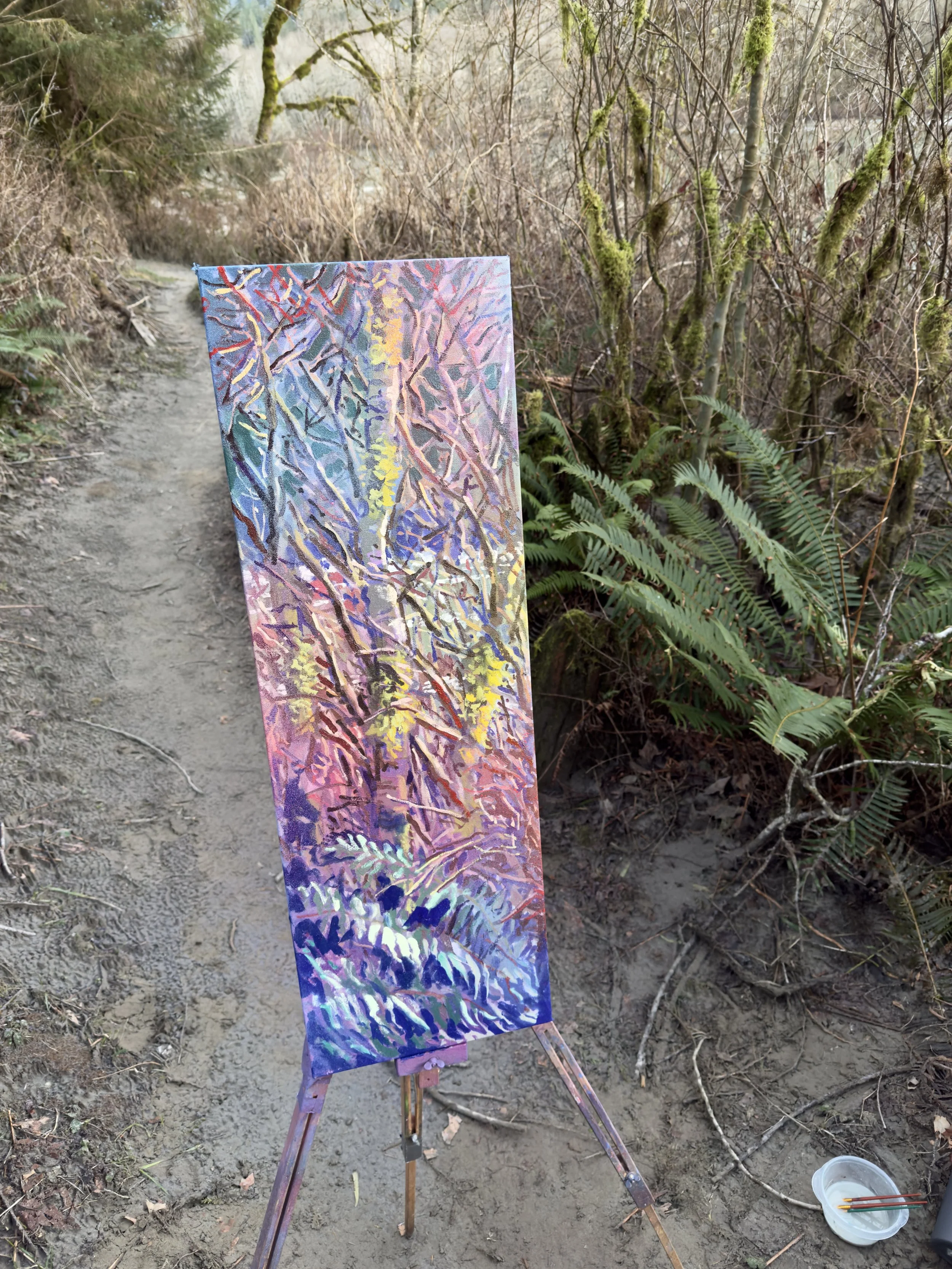 An easel with an impressionist-style painting of a tree with branches and foliage, set outdoors on a dirt path surrounded by leafless trees and green ferns.