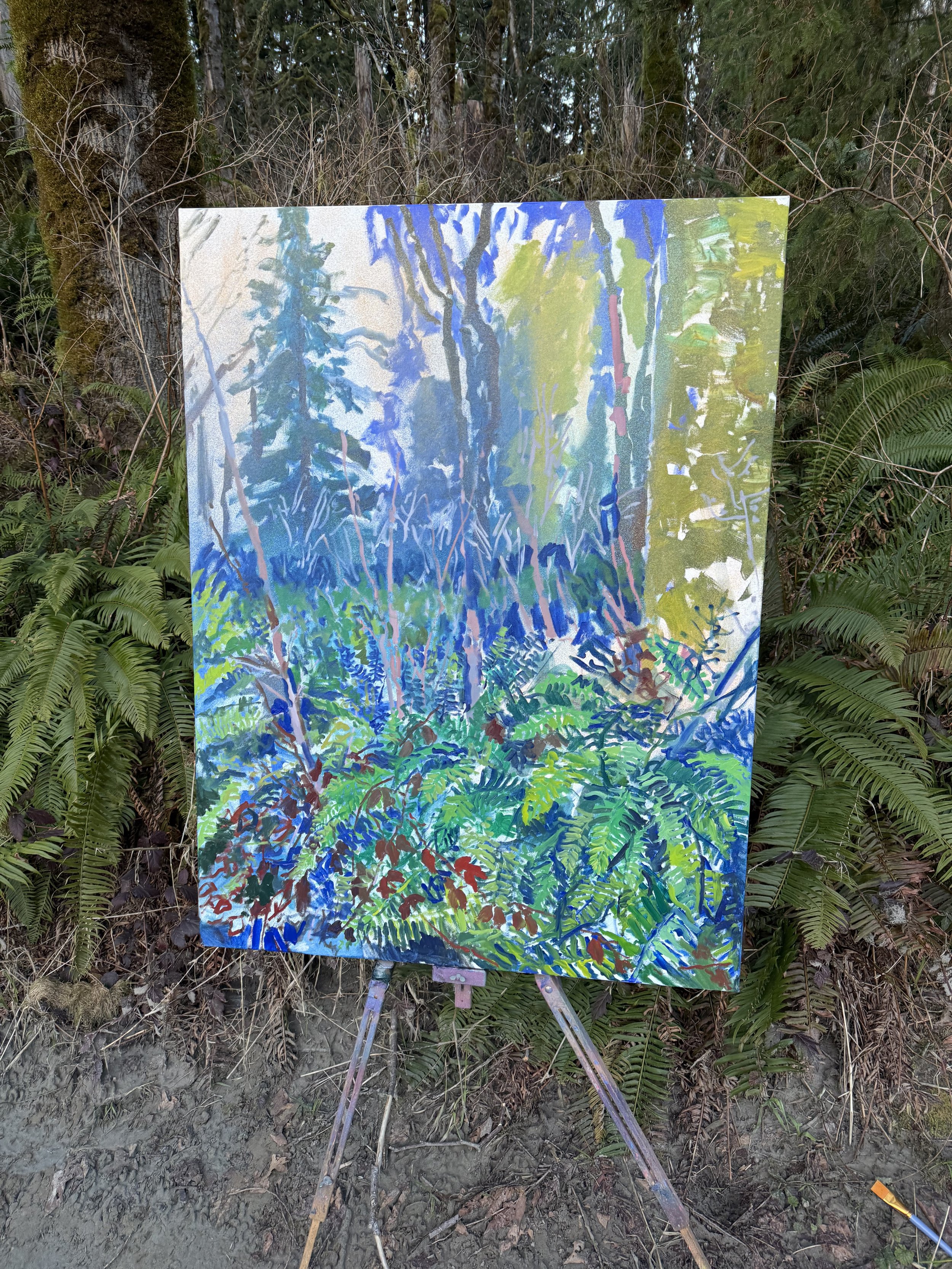 An outdoor painting on canvas of a forest scene with trees and foliage, mounted on an easel in a wooded area with ferns and trees in the background.