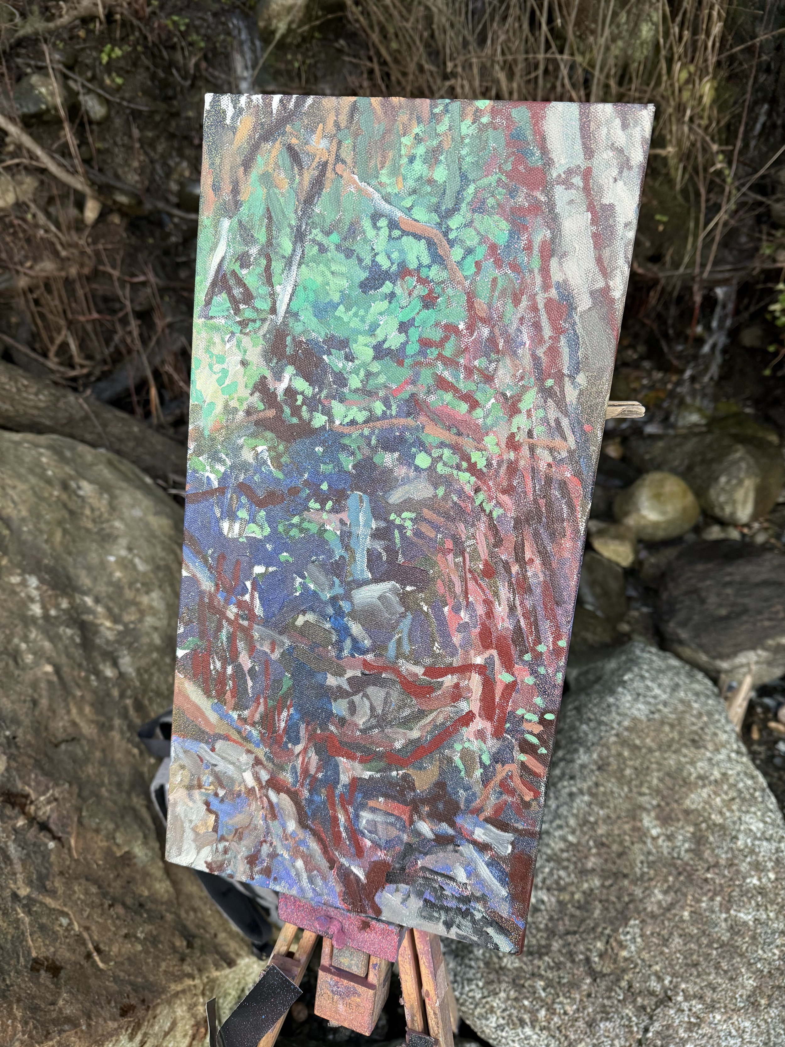 An impressionistic painting of a snowy mountain landscape with trees and rocks, on an easel outdoors among rocks and bushes.