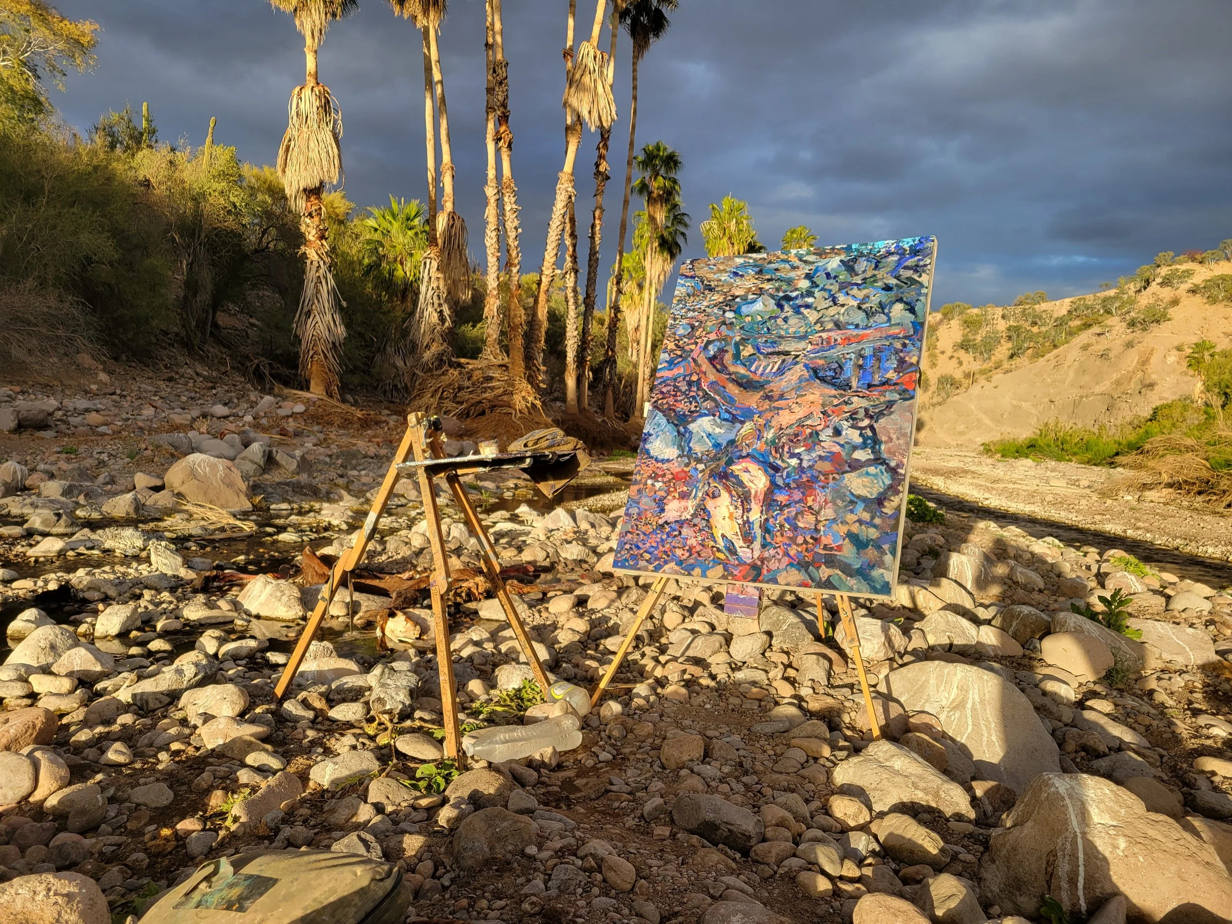 An artist's painting set up outdoors on rocky ground, with a table holding art supplies, a canvas of a colorful landscape, and palm trees in the background under a cloudy sky during late afternoon or early evening.