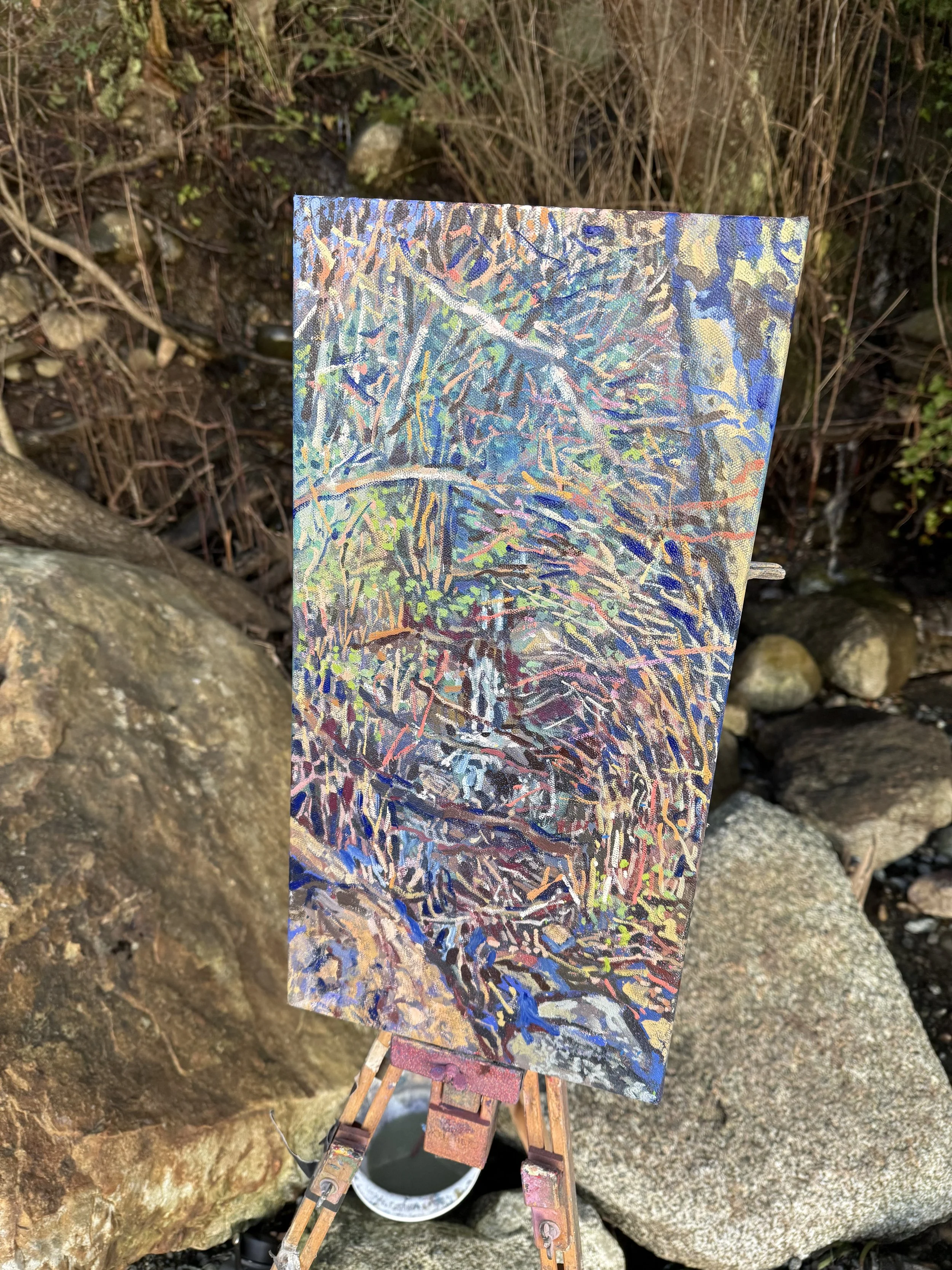 An abstract painting featuring a dense array of colorful lines and plant-like shapes, placed on a small easel among rocks and outdoor greenery.