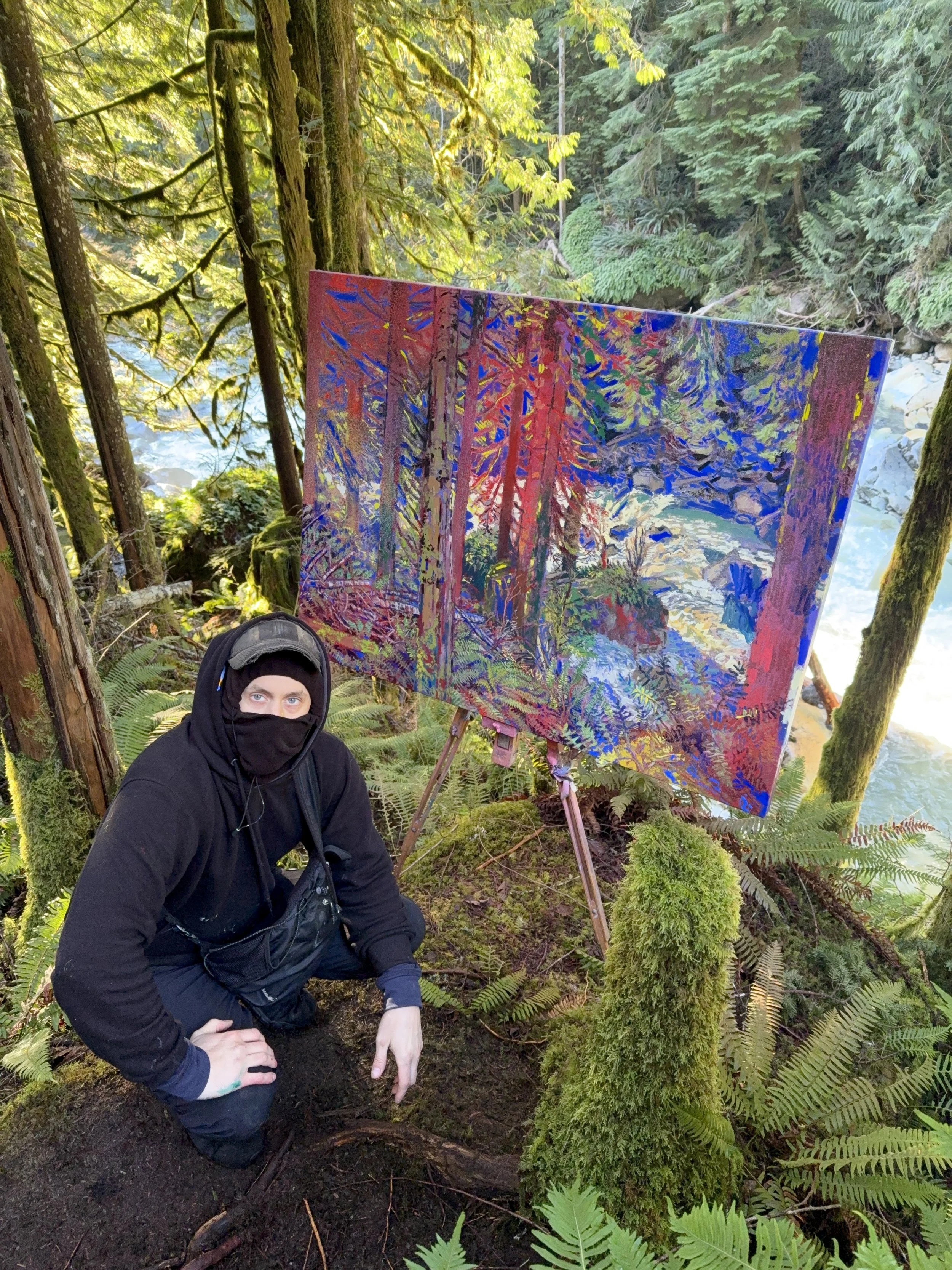Person dressed in black with a face covering, kneeling in a forest next to a colorful painting of a forest scene on an easel.