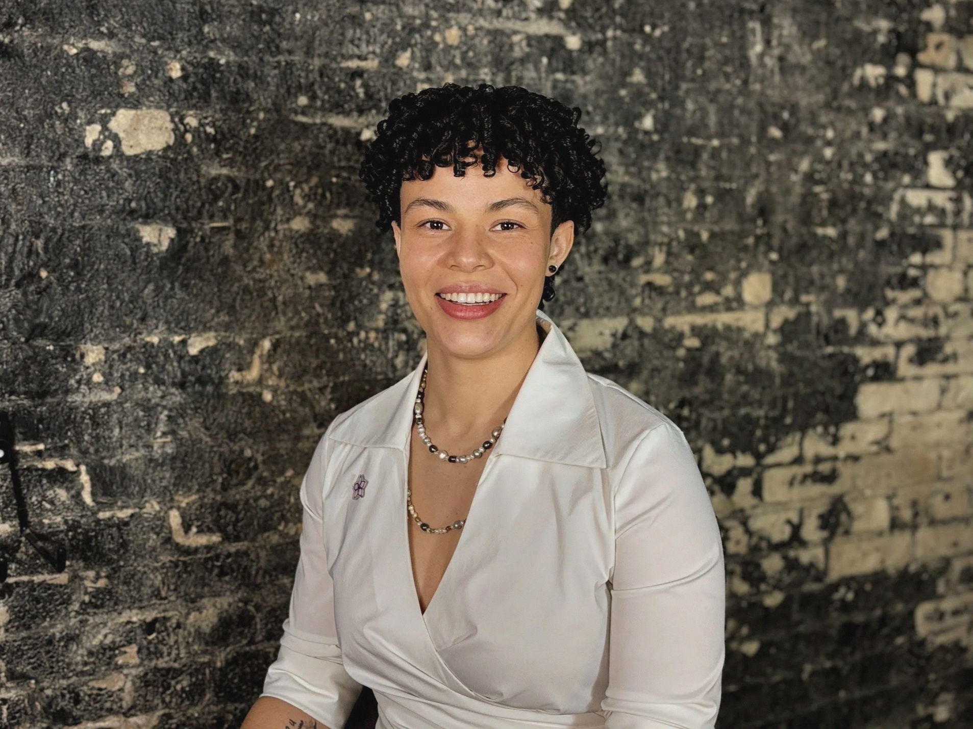 A person with short, curly black hair sitting against a grey background, wearing a sleeveless maroon top and dark jeans, looking at the camera with a slight smile.