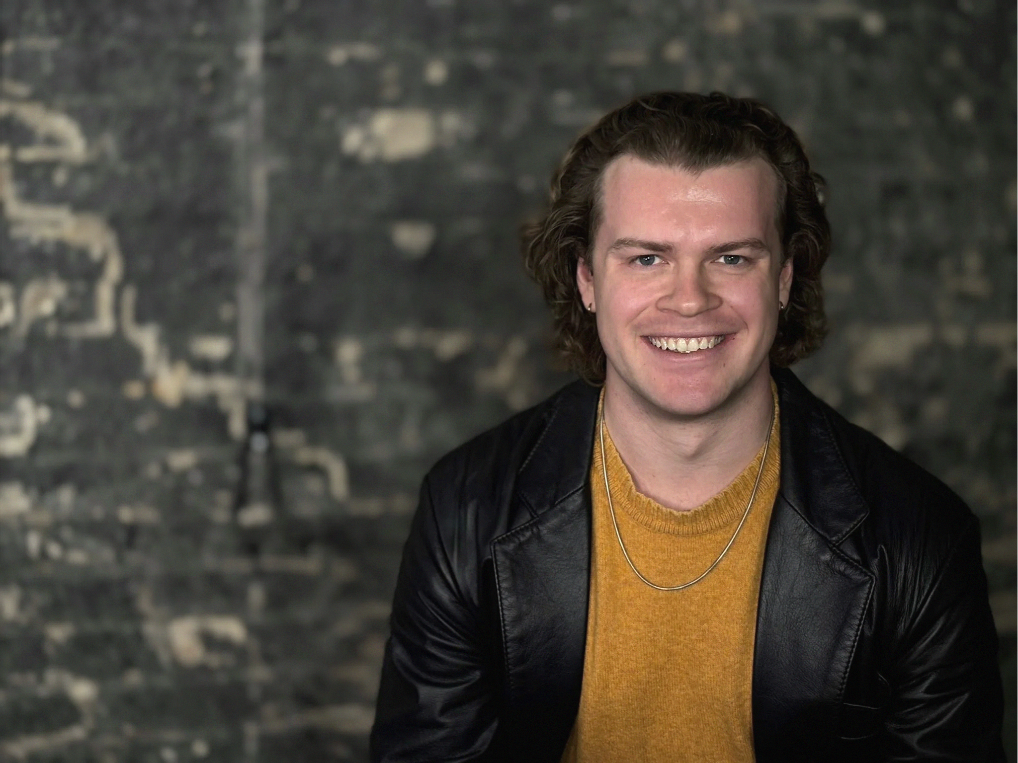 Smiling man with curly hair and earrings wearing a yellow sweater and black leather jacket, standing in front of a textured dark background.