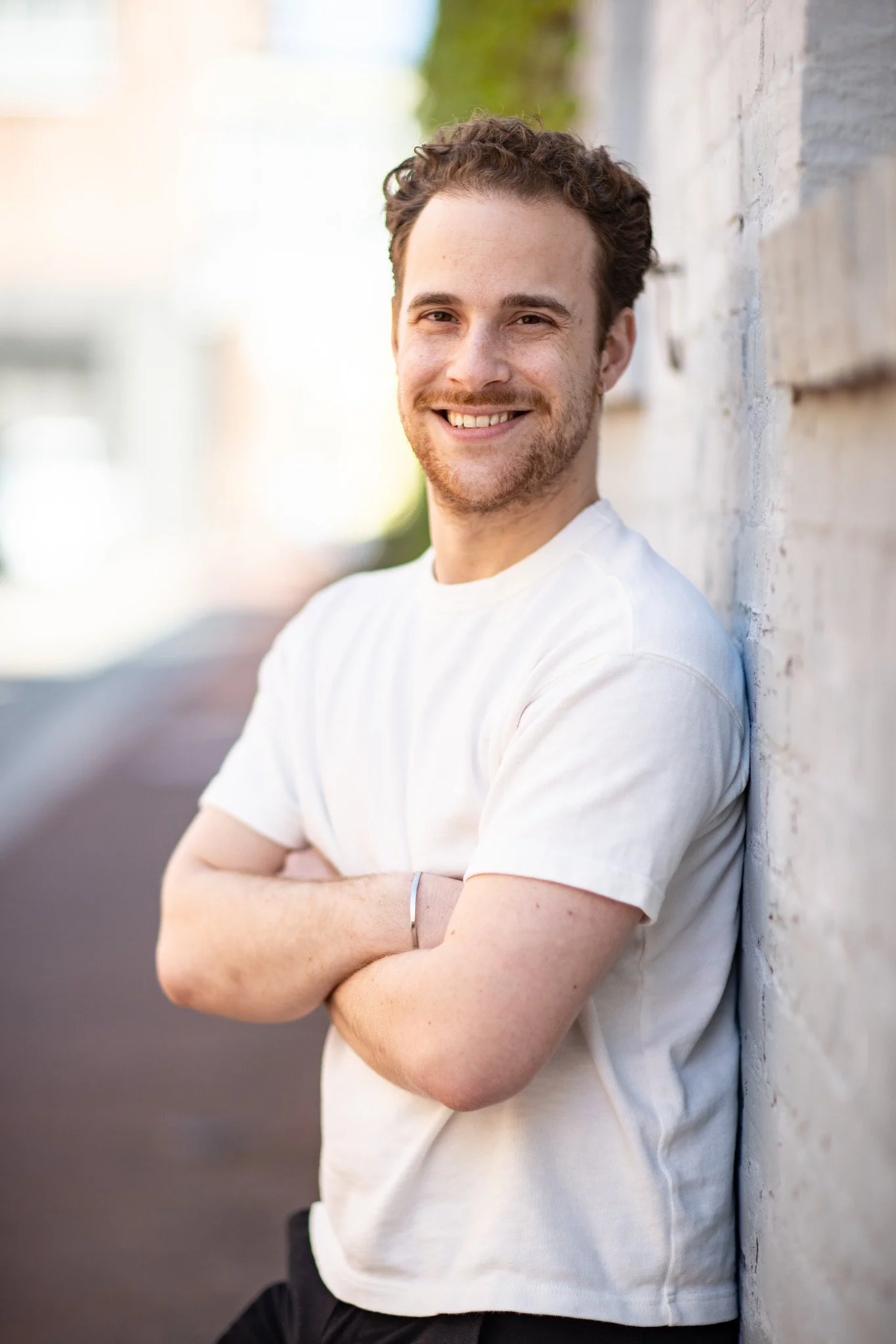 A young man with light brown hair and a beard, wearing a dark suit, white shirt, and dark tie, smiling while standing in front of a brick wall.