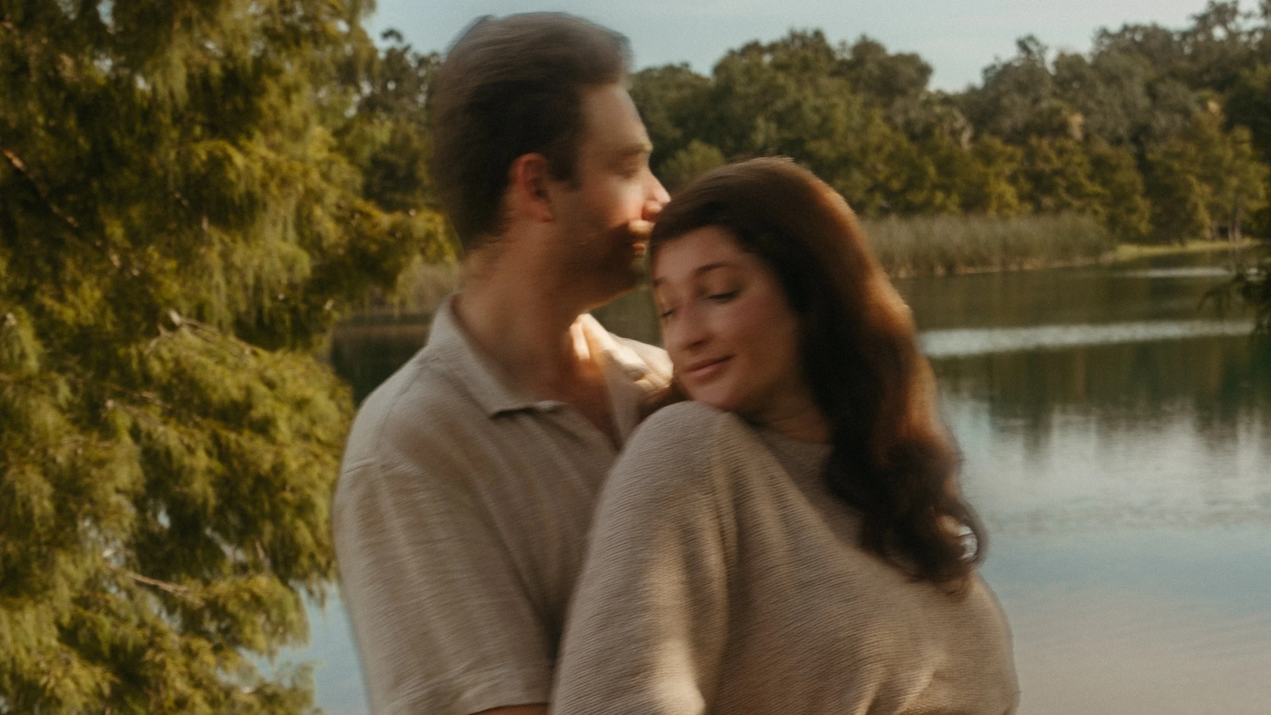 A couple embracing by a lake surrounded by trees and greenery, with clear weather.